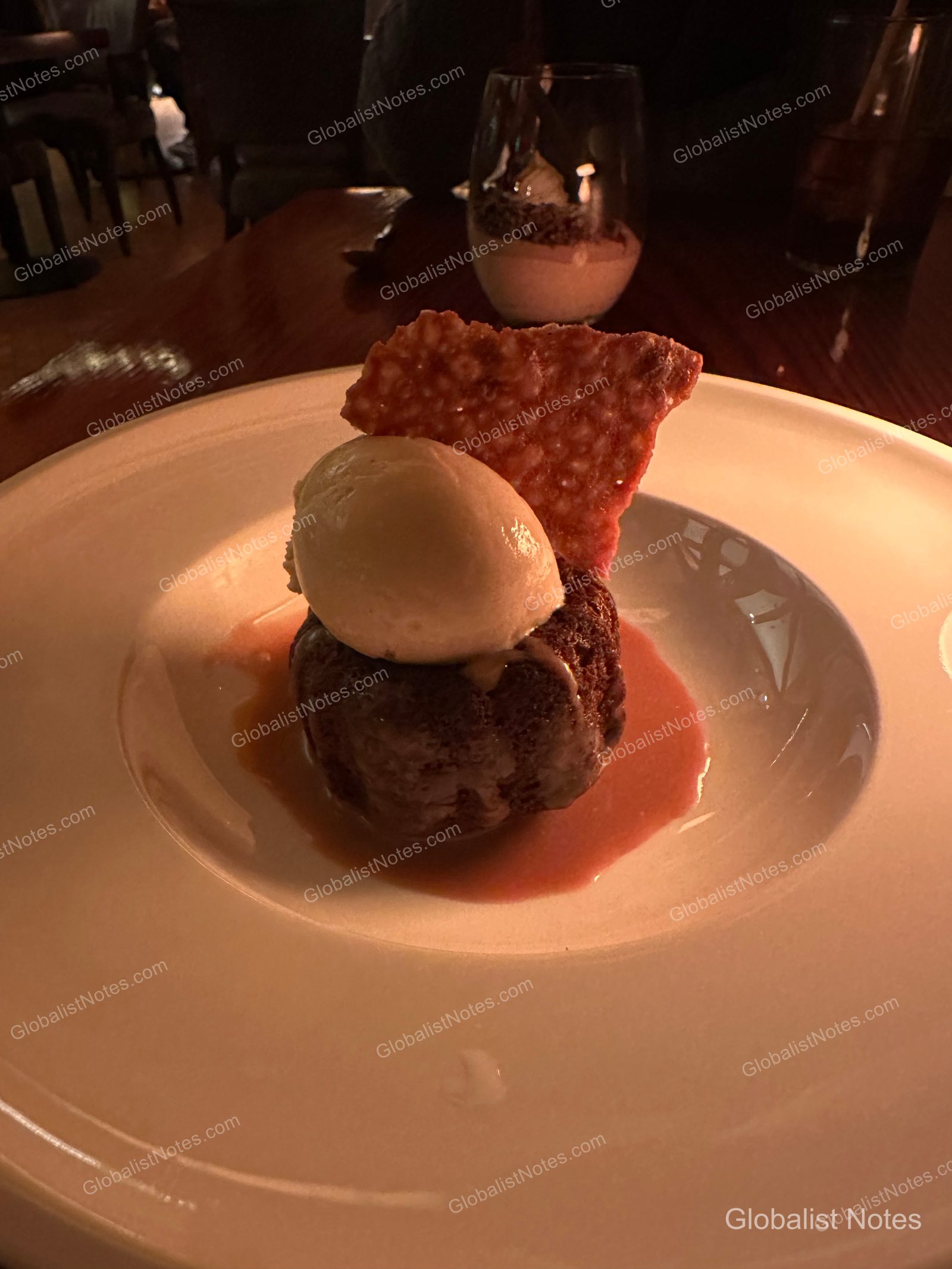 A dessert plate on a table at the Alila Ventana Big Sur restaurant in dim lighting. It features a warm cake topped with a scoop of vanilla ice cream and a crispy tuile, served over a red sauce.