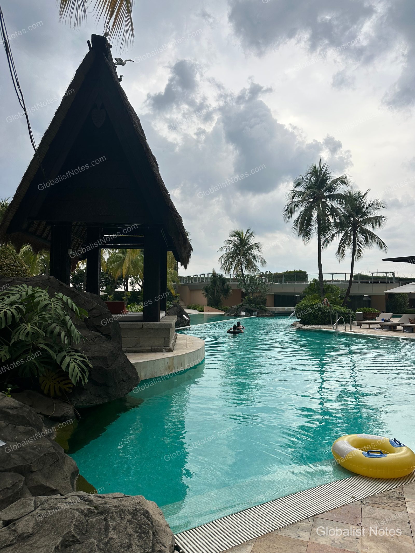View of the outdoor pool at Grand Hyatt Jakarta, Indonesia. Guests are swimming in the pool, which features thatched-roof gazebos, rock formations, and palm trees; a yellow pool float is visible in the water.