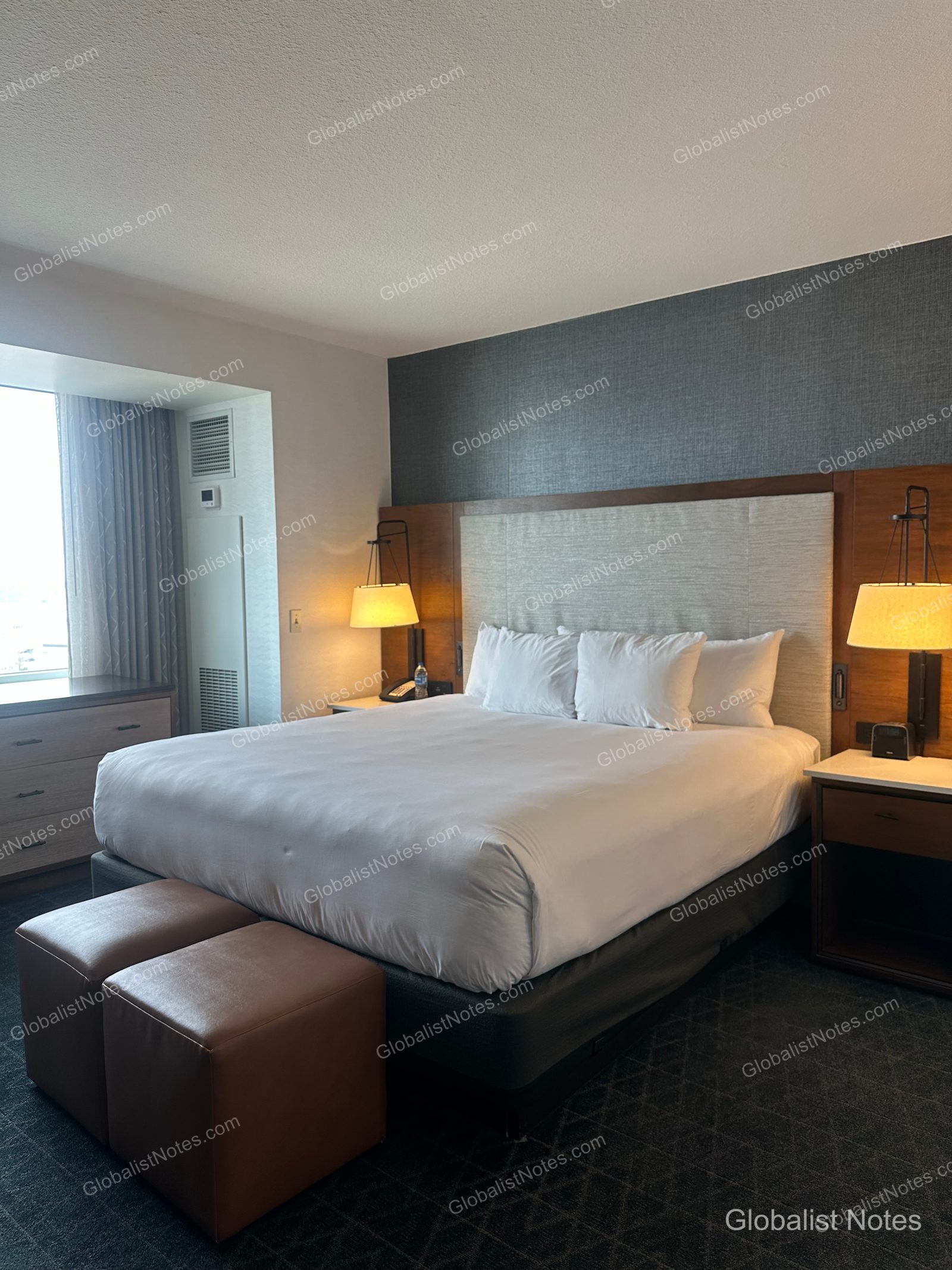 Bedroom of the Executive Suite at Grand Hyatt San Antonio River Walk. The king bed features a headboard with integrated lighting, two brown leather stools at the foot of the bed, and a dresser positioned by the window.