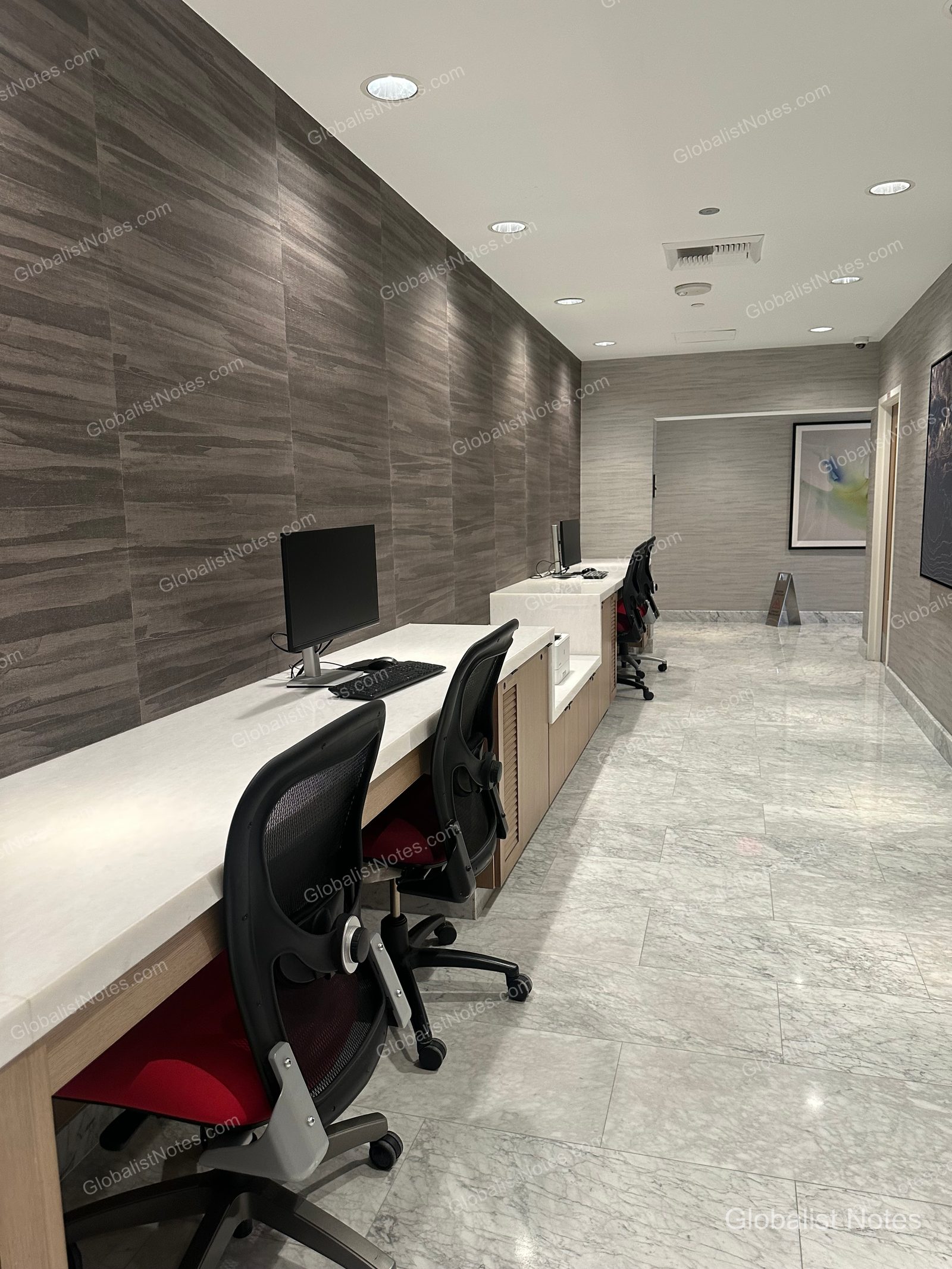 Hyatt Regency Newport Beach West 商務中心，長型白色桌面上擺放電腦與鍵盤，配有黑色網狀辦公椅。牆面採用深灰色木質裝飾，地面為淺色大理石磁磚。