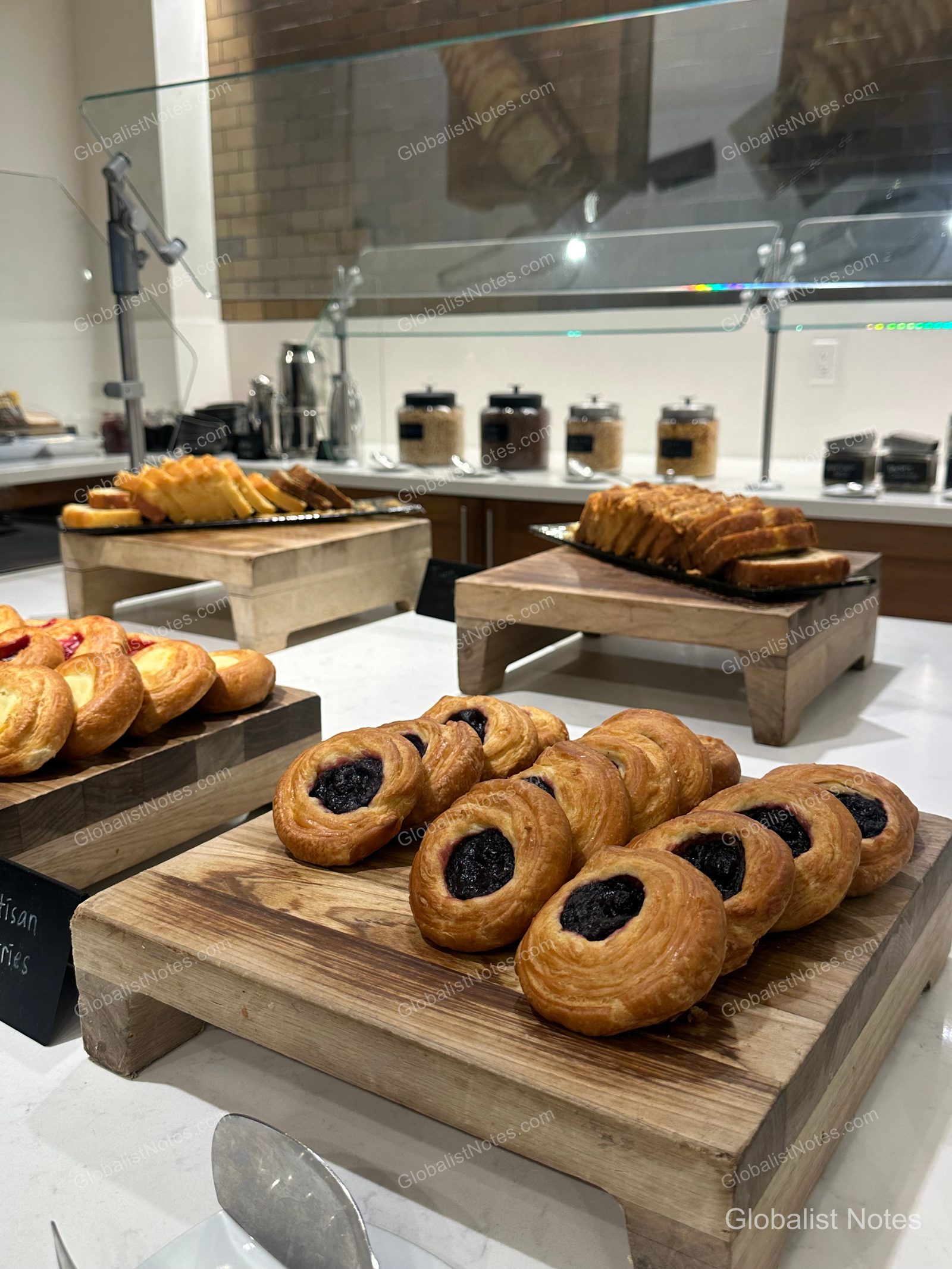 A selection of pastries, including blueberry danishes and sliced bread, neatly displayed on wooden stands at the Hyatt Regency Los Angeles International Airport breakfast buffet.