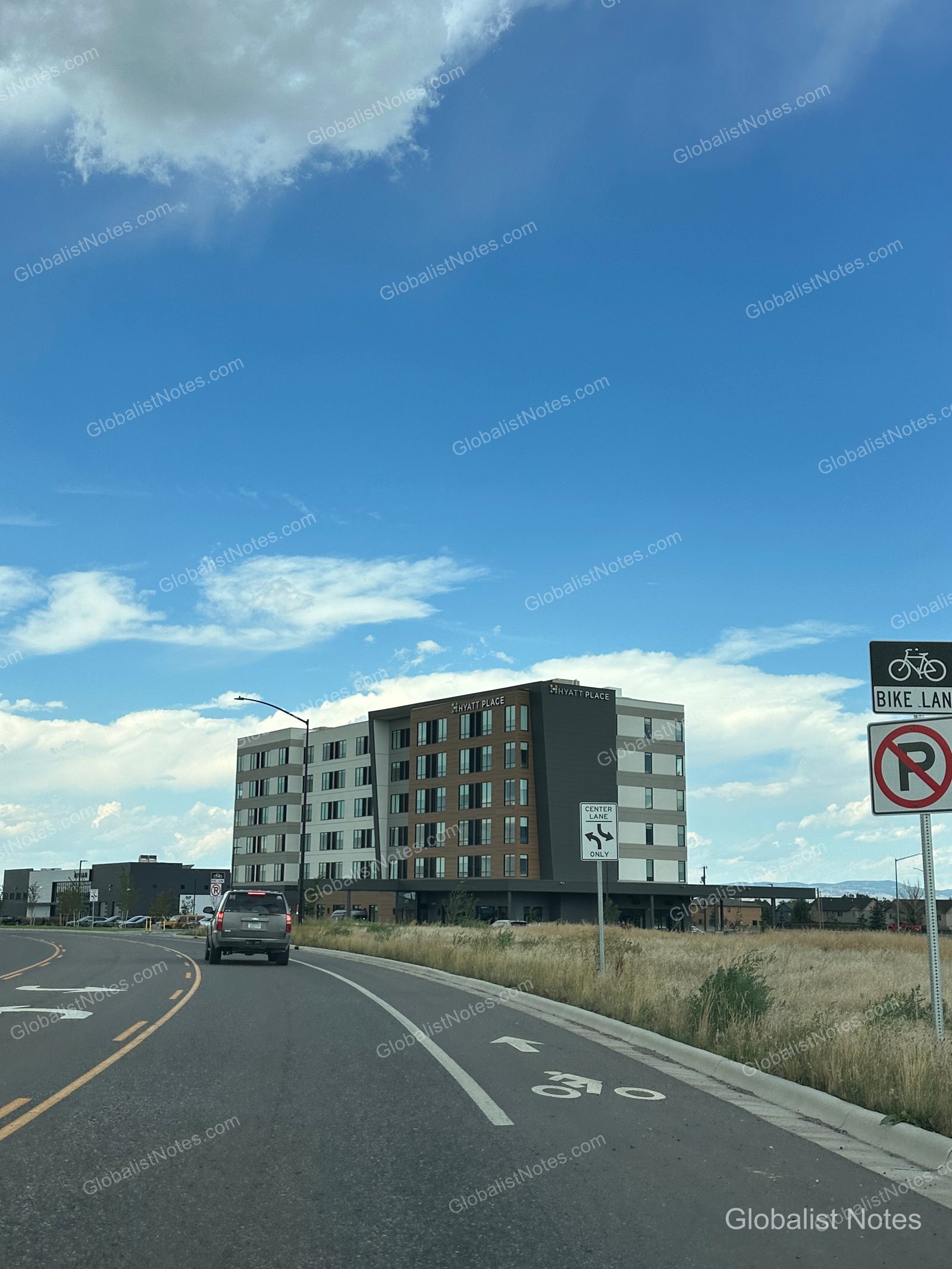 米国ベルグレードにあるhyatt place Bozeman Yellowstone Airportの外観。モダンな建物と周辺の草地、自転車道の標識、晴天の下をホテルへ向かう車が見える。