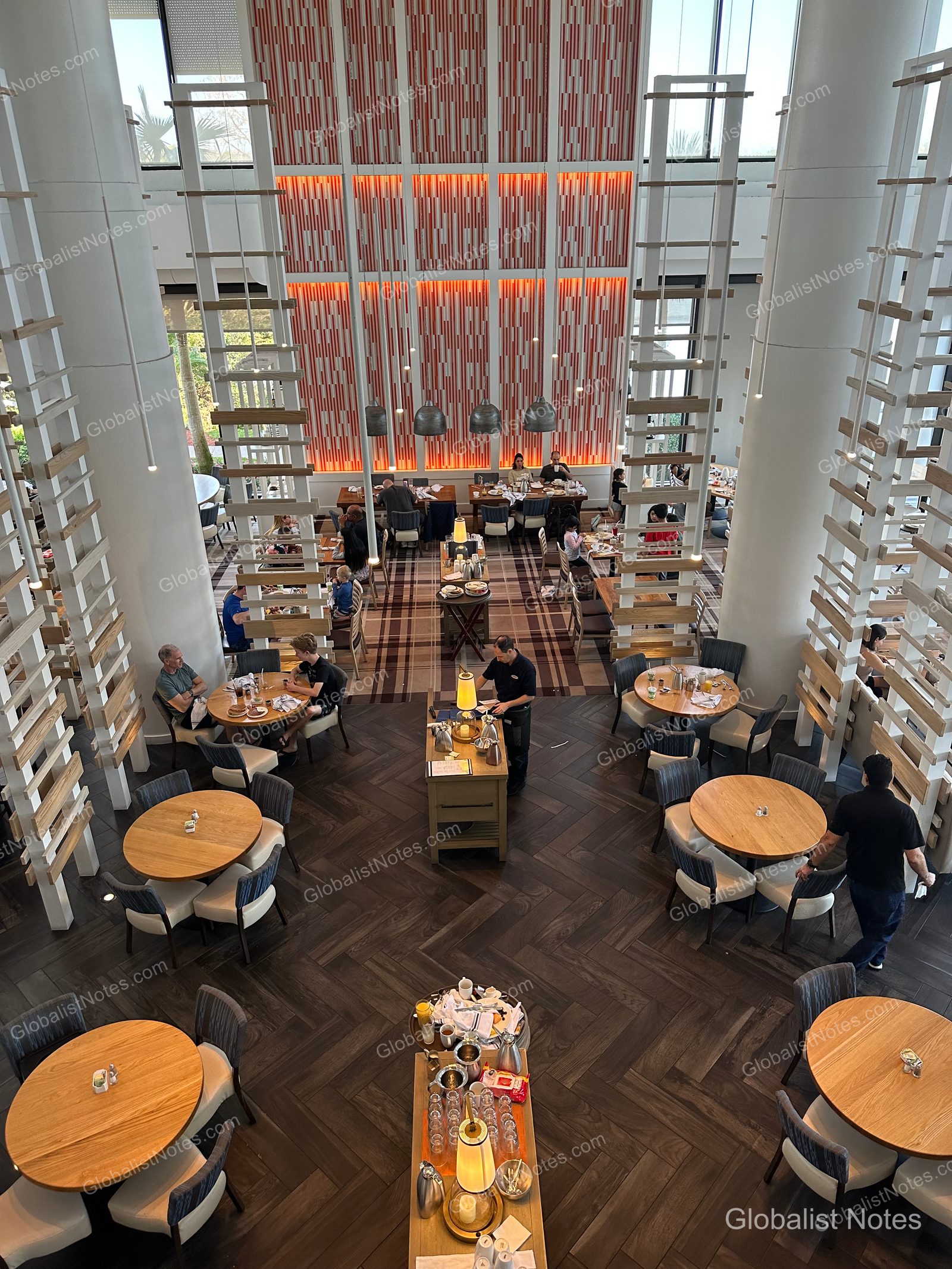 Overhead view of the high-ceilinged, open-plan dining space at Hyatt Regency Grand Cypress. Light wood round tables and chairs are set on a herringbone-patterned floor, with a service cart and tableware in the center.