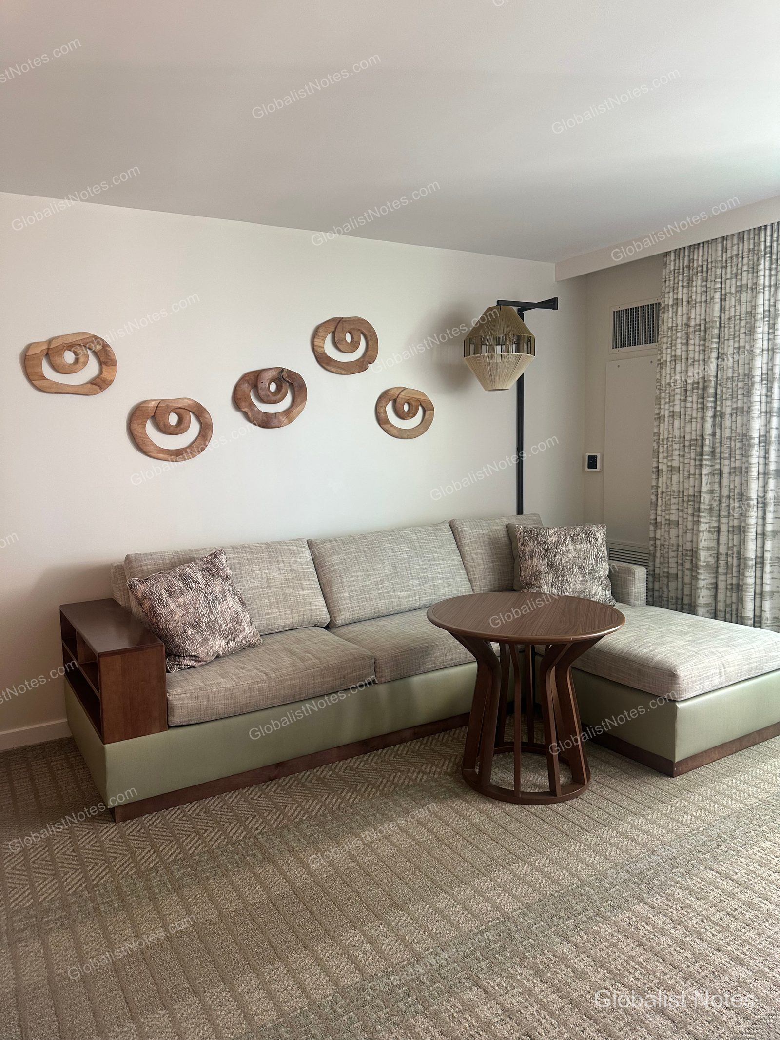 Interior view of a standard king room at Hyatt Regency Maui Resort and Spa. The space features a beige sofa, a round wooden table, and five spiral wooden wall decorations next to a uniquely designed lamp.
