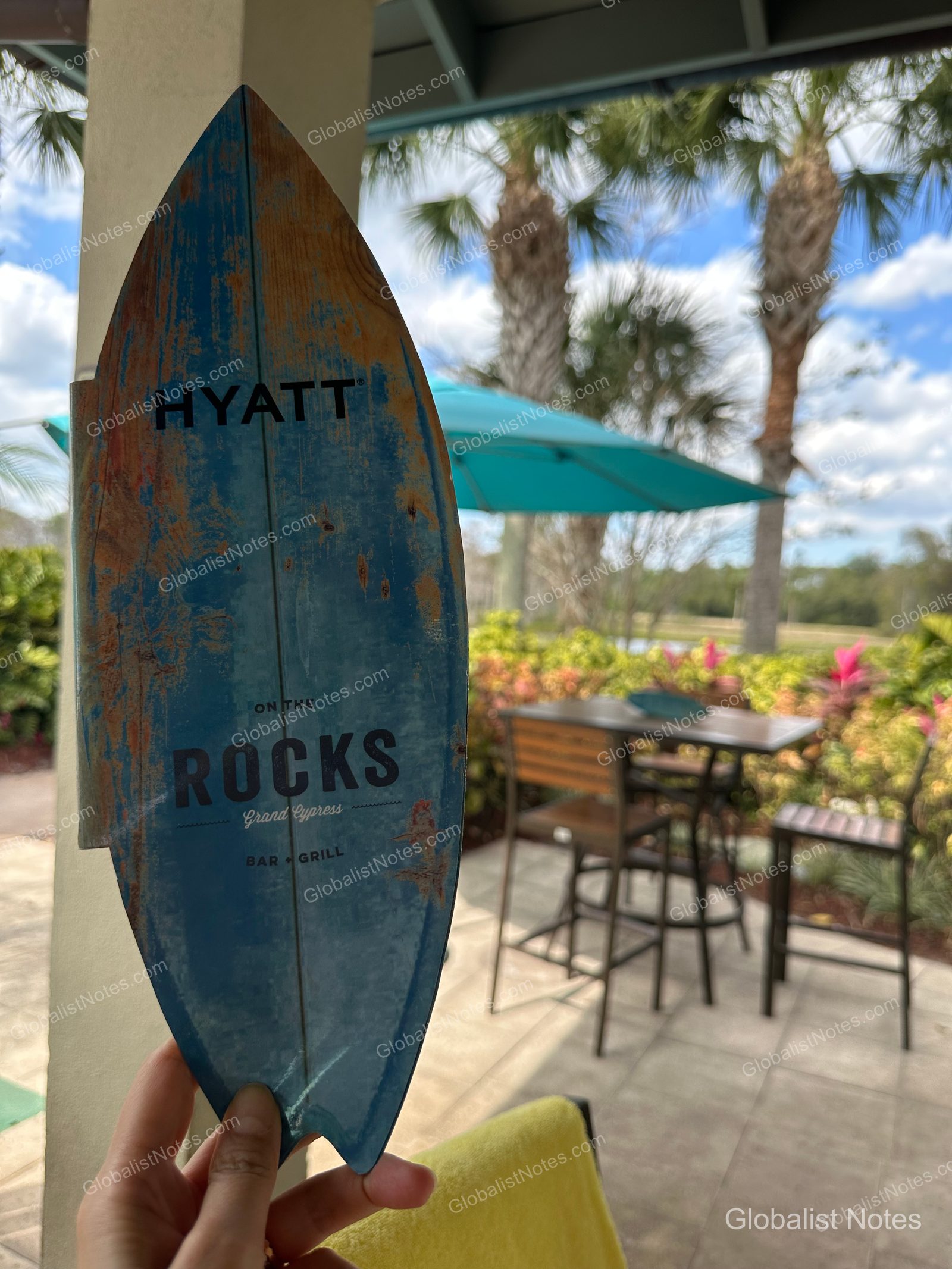 A hand holding a surfboard-shaped sign with the 'On the Rocks' Bar & Grill logo at Hyatt Regency Grand Cypress, with outdoor terrace seating, umbrellas, and palm trees in the background.
