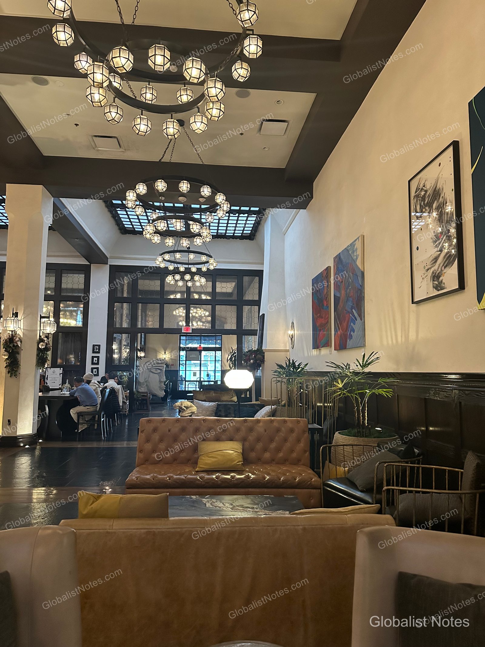 View of the lobby lounge at Hotel Figueroa in Los Angeles. Leather sofas and tables are arranged beneath several circular chandeliers hanging from the high ceiling, with abstract art on the walls and a view of the breakfast area and entrance in the background.