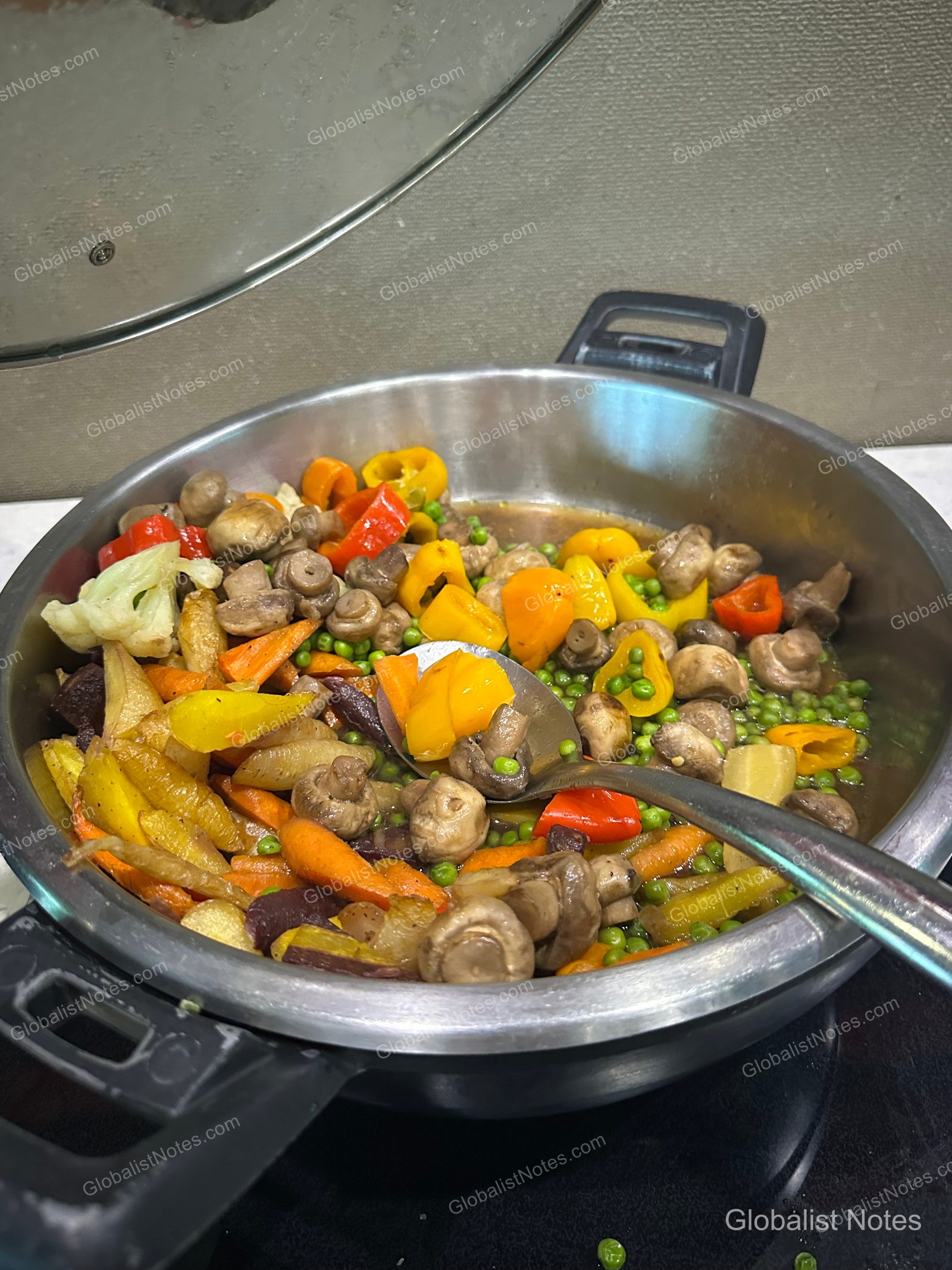 Warm vegetable stir-fry served in a stainless steel chafing dish at the Hyatt Regency Vancouver breakfast buffet. The dish includes a mix of mushrooms, yellow and red bell peppers, carrots, peas, and cauliflower, with a metal serving spoon resting in the center.