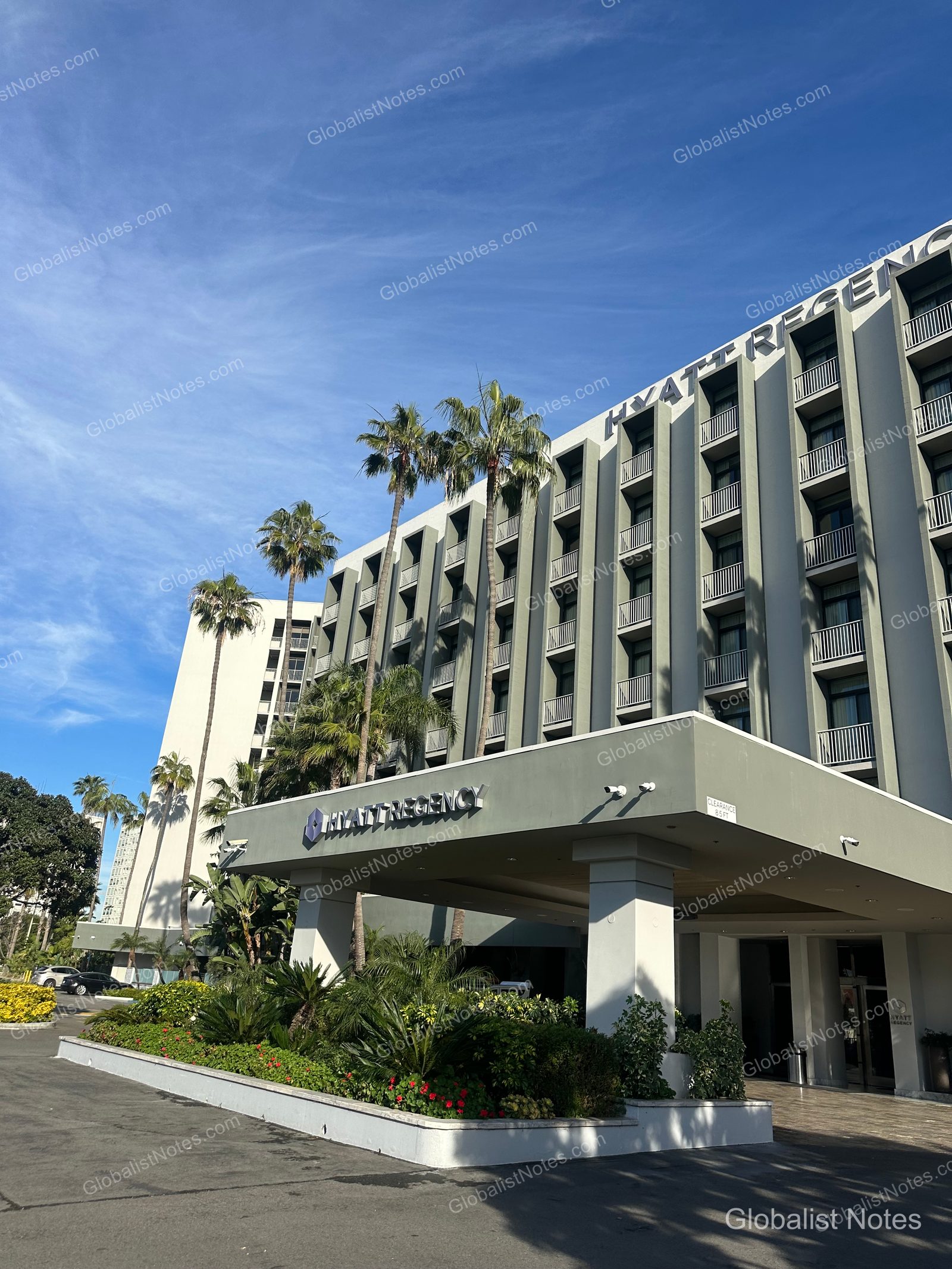 位於加州 Newport Beach 的 Hyatt Regency 外觀，高大棕櫚樹整齊排列，飯店入口雨遮與帶有陽台的客房建築映襯在藍天下。