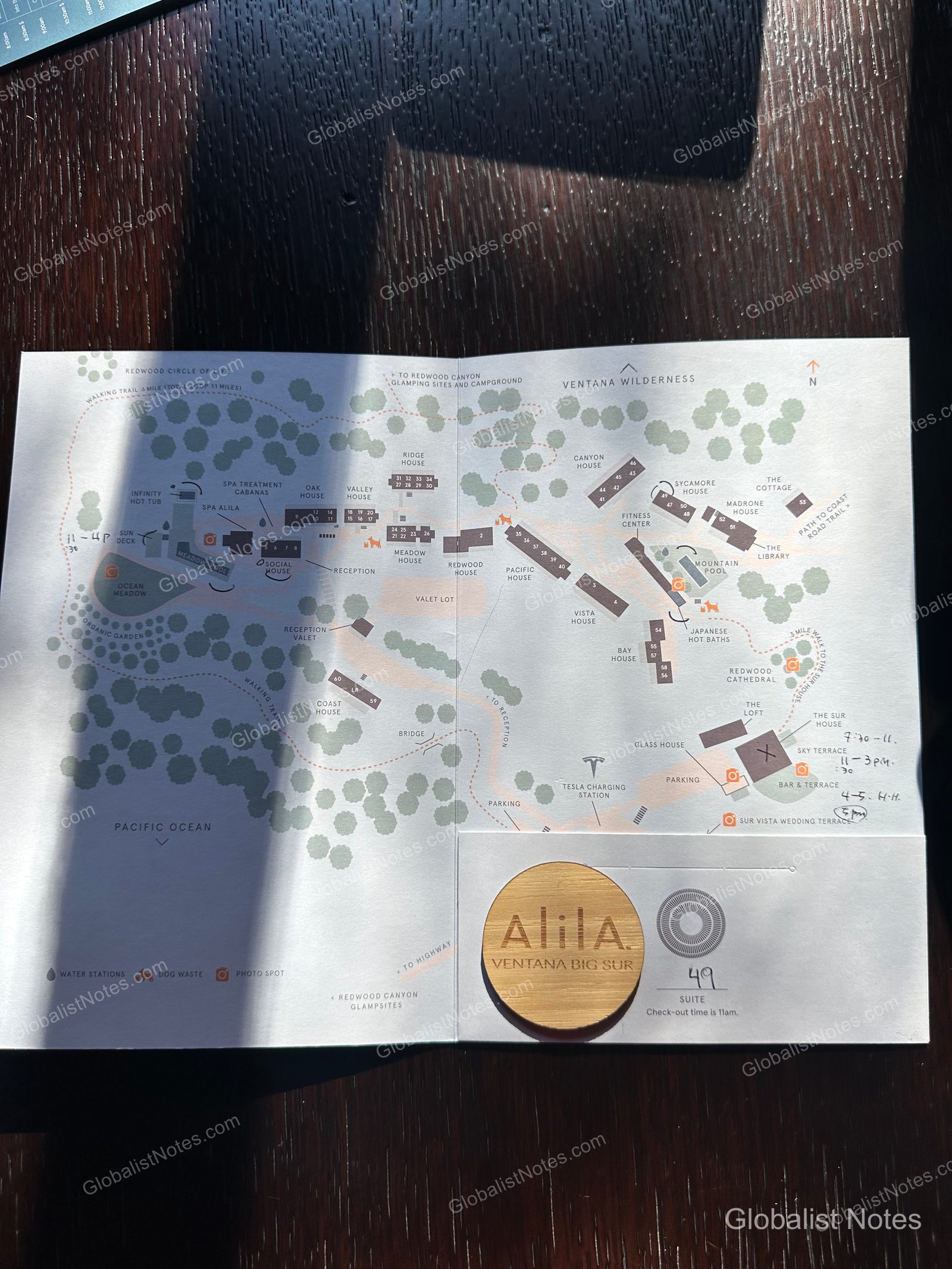 A resort map of Alila Ventana Big Sur showing the reception, guest room buildings, infinity pool, Japanese hot baths, and walking trails. A wooden key card for Junior Suite 49, with the checkout time written on it, is placed at the bottom.