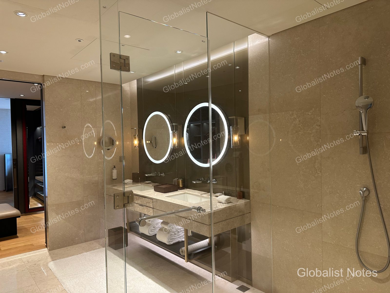 Bathroom in a One Bedroom Suite at Grand Hyatt Jeju. The space features marble walls and flooring, a double vanity with circular LED-lit mirrors, and a glass-enclosed shower. A hallway leading to the main room is visible on the left.