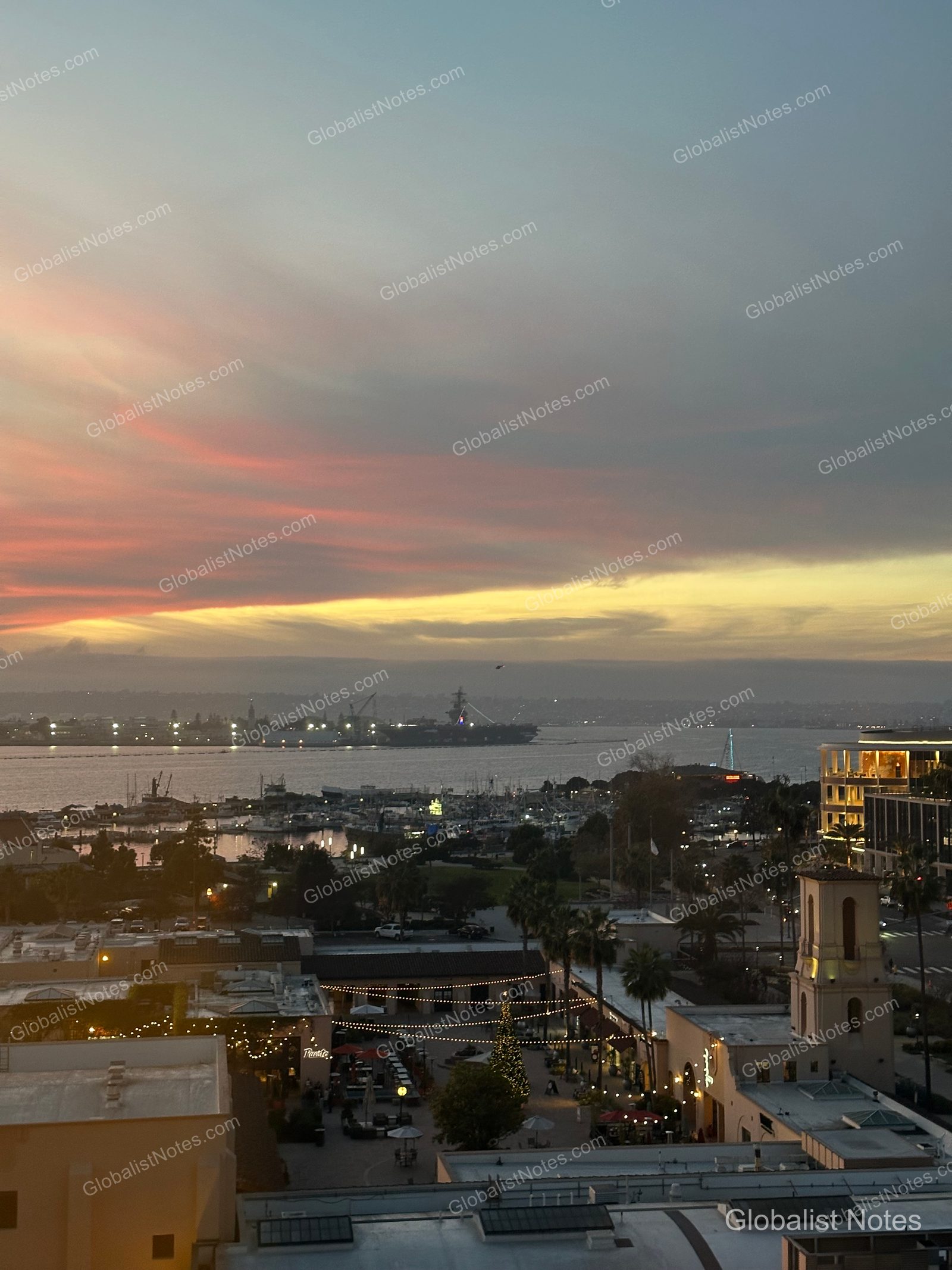 從 Manchester Grand Hyatt San Diego 客房俯瞰的黃昏景色。在粉色與黃色交織的晚霞下，港口停泊著船隻，遠處可見軍艦，下方的廣場則點亮了聖誕燈飾。