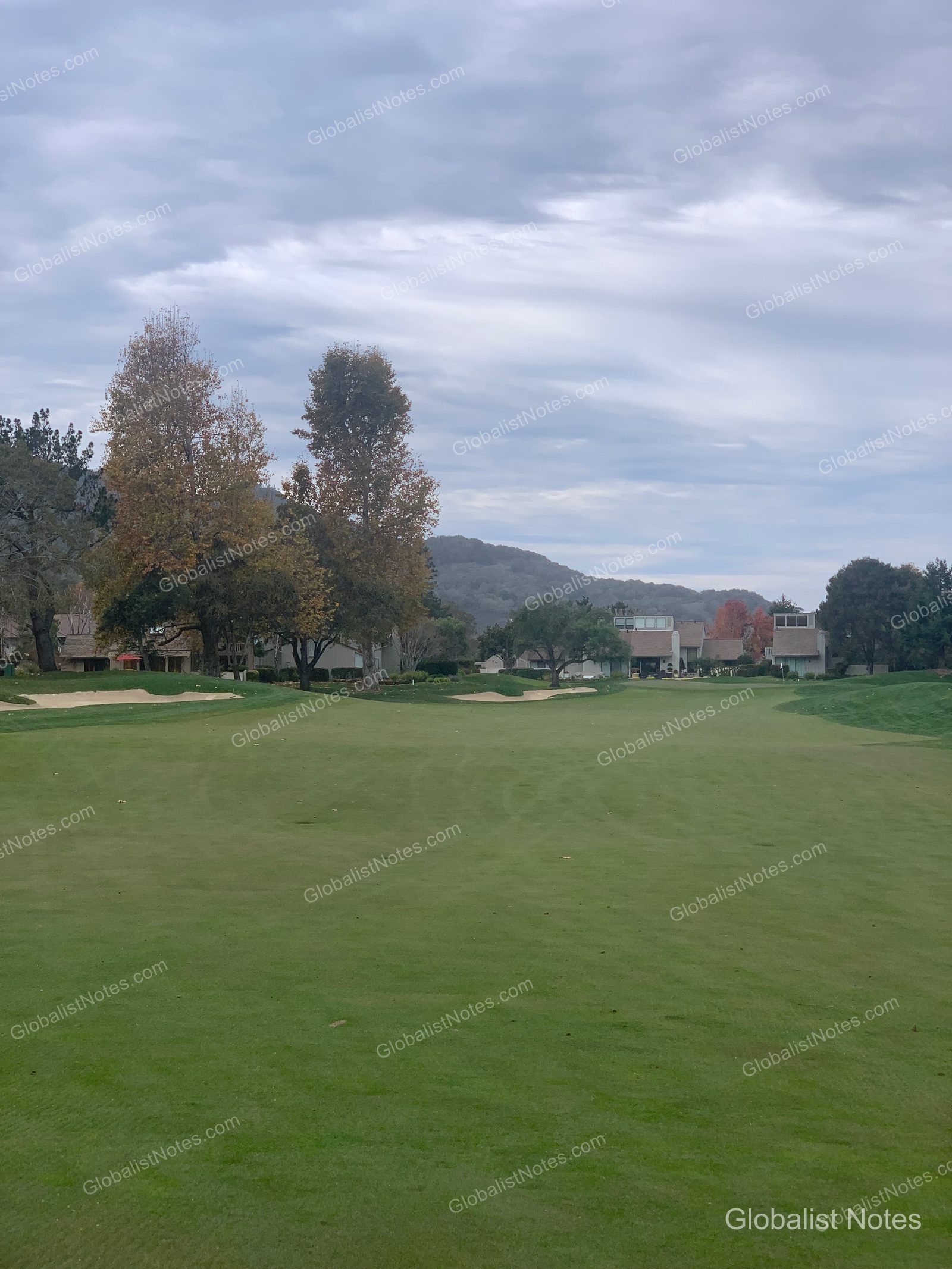 Carmel Valley Ranch - The Unbound Collection by Hyattのゴルフコース全景。フェアウェイの奥にバンカーと木々が続き、遠くには山々とリゾートの建物が曇り空の下に広がっています。