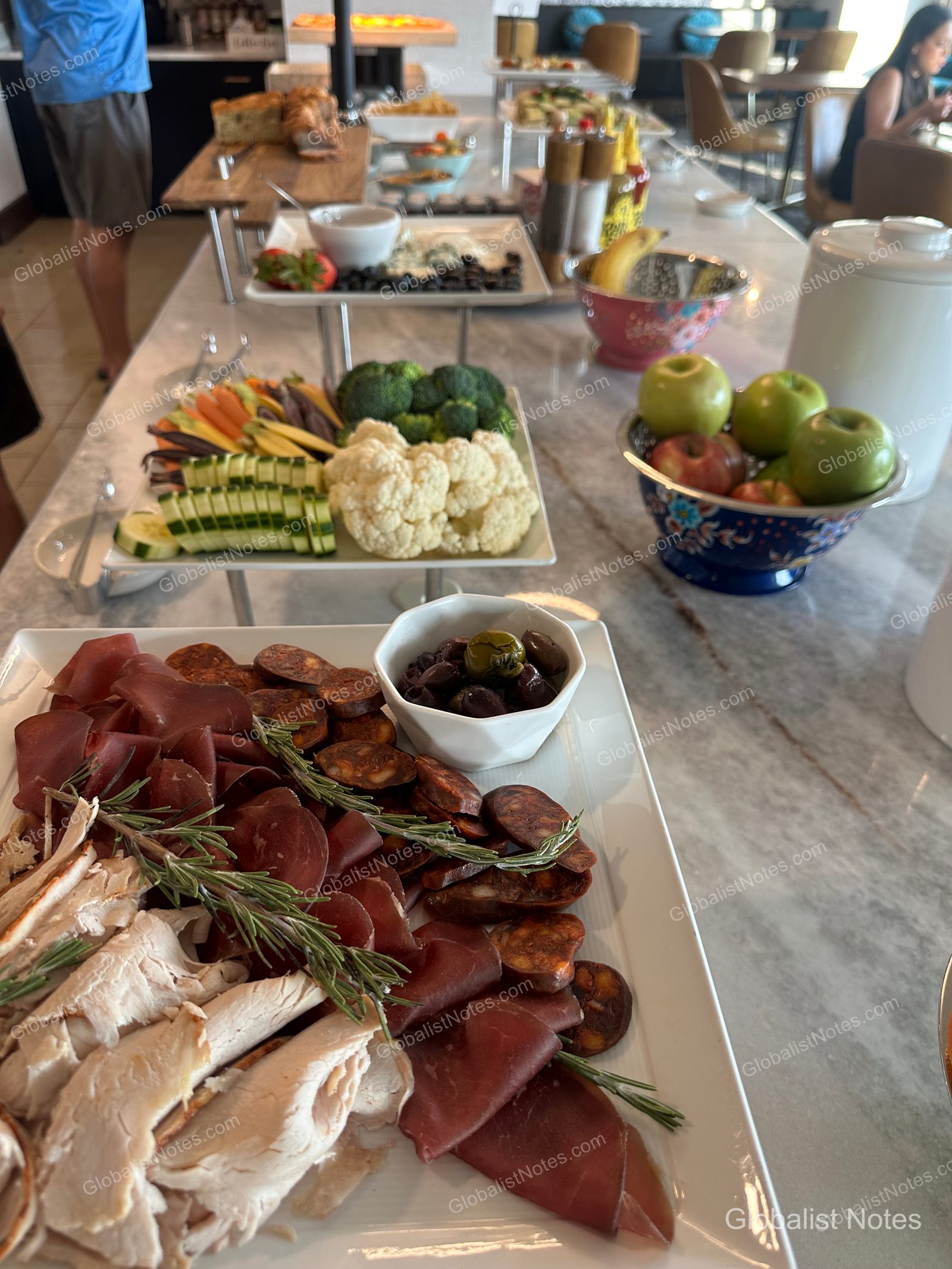 Buffet spread in the Grand Hyatt San Antonio River Walk lounge, featuring plates of sliced ham, chicken, and olives in the foreground, with bowls of fresh broccoli, cauliflower, cucumber, carrots, and apples arranged on a marble table.