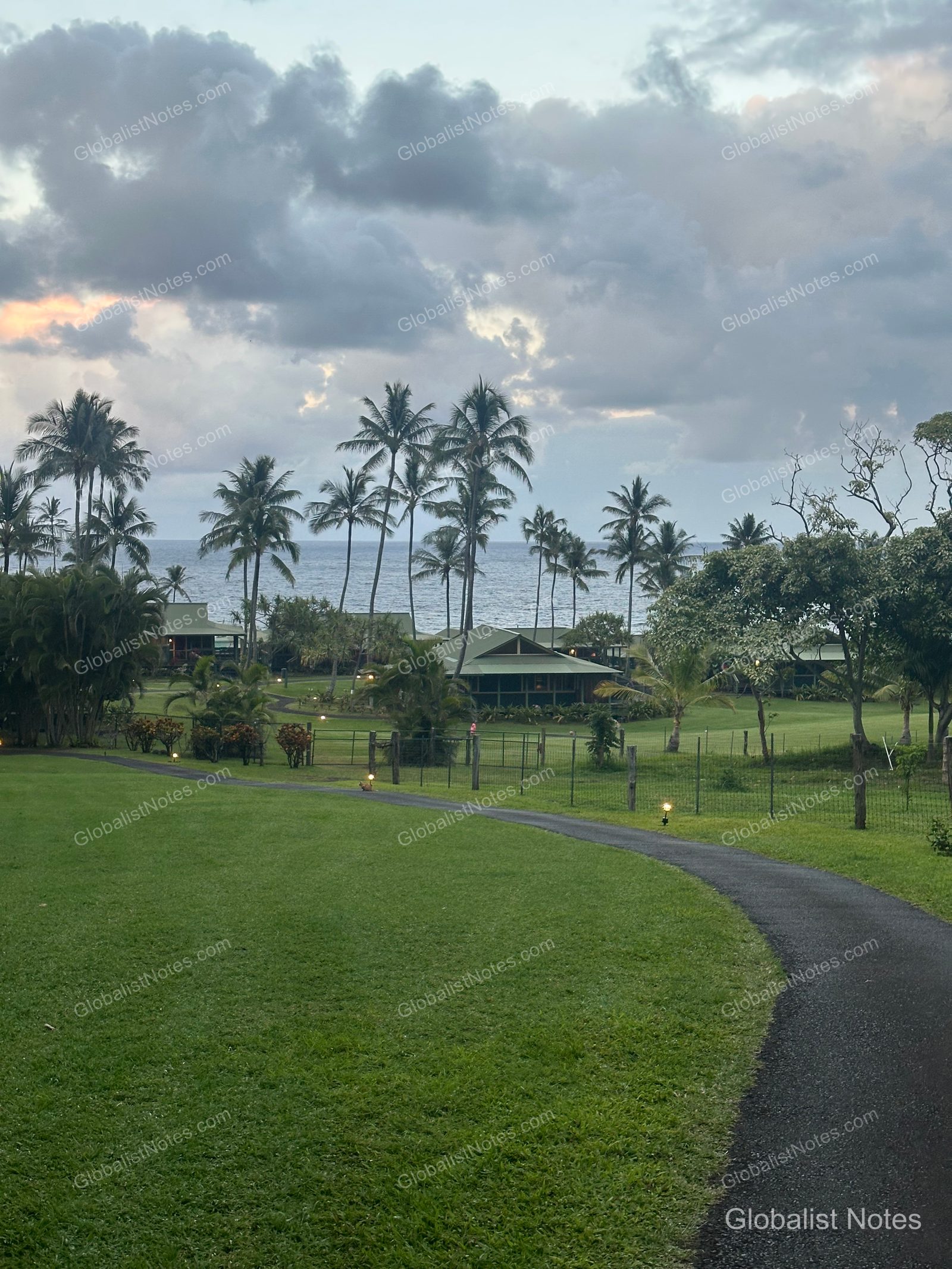 A view of Hana-Maui Resort featuring a gently curving walkway and expansive lawns, with palm trees and the Pacific Ocean in the background. The resort's low-rise buildings are set against a cloudy sky.