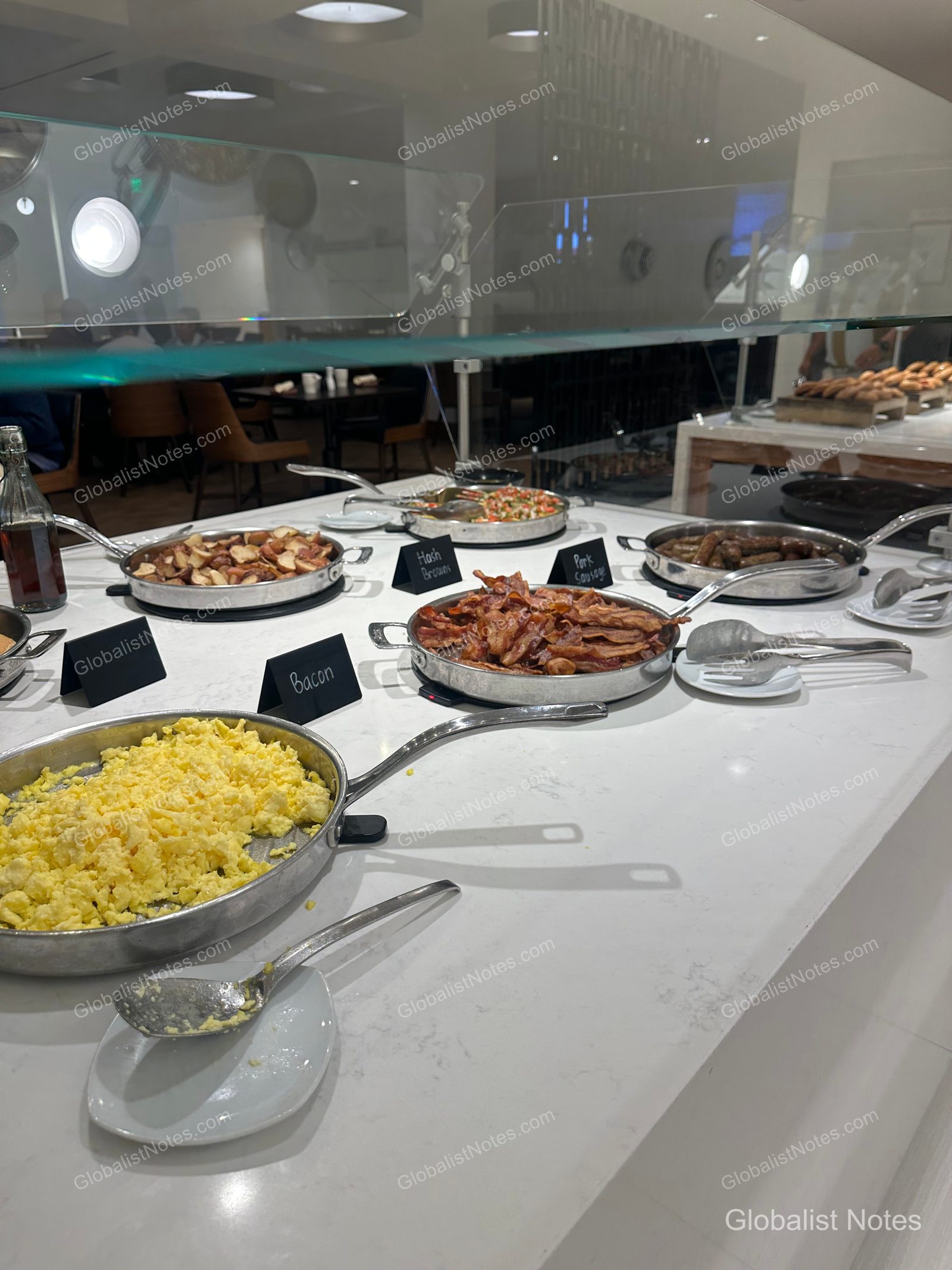 The hot food station at the Hyatt Regency Los Angeles International Airport breakfast buffet. Stainless steel chafing dishes containing scrambled eggs, bacon, hash browns, and sausages are arranged on a white marble countertop, with black labels identifying each dish.