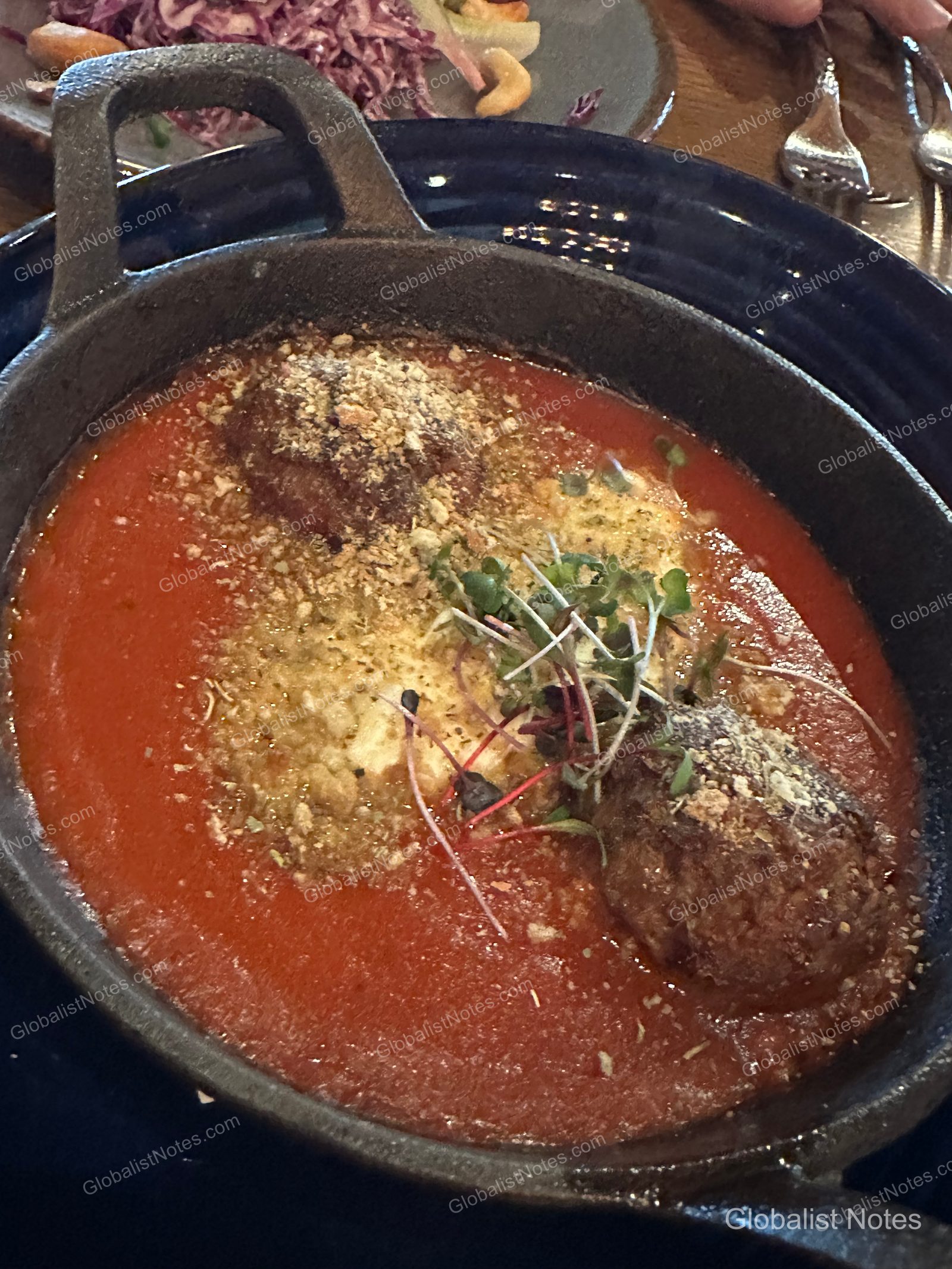 미라발 오스틴 리조트 앤 스파의 저녁 식사로 나온 무쇠 팬 요리입니다. 붉은 토마토소스 위에 두 개의 미트볼이 놓여 있고, 그 위로 치즈 가루와 신선한 새싹 채소가 올려져 있습니다. 배경에는 샐러드 접시와 은색 식기류가 일부 보입니다.