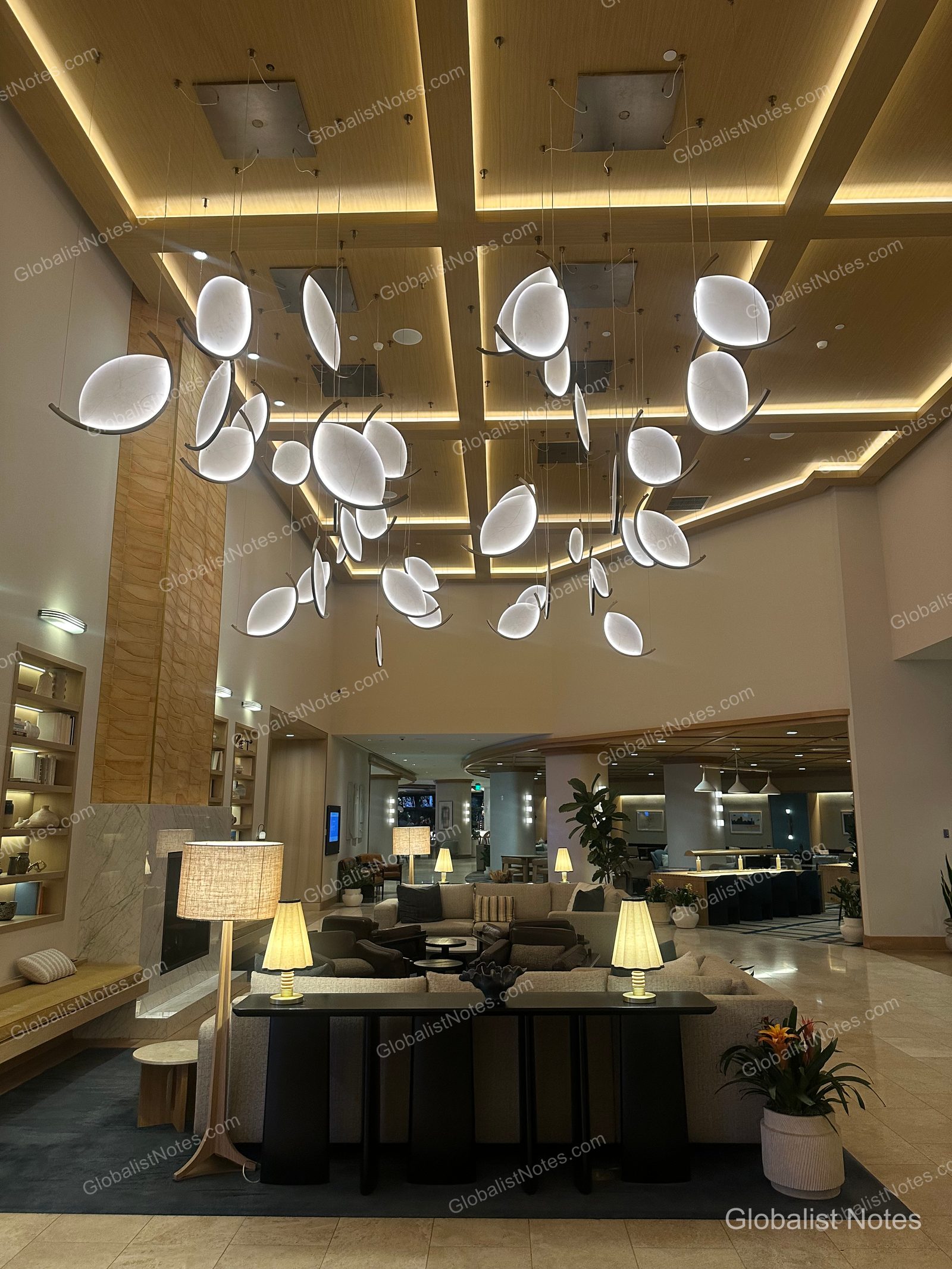 Hyatt Regency Irvine 大廳挑高天花板懸掛著葉片造型燈飾，下方規劃了寬敞的休憩區，配置沙發與茶几。牆面以層架與壁爐裝飾，整體空間呈現溫暖沉穩的氛圍。