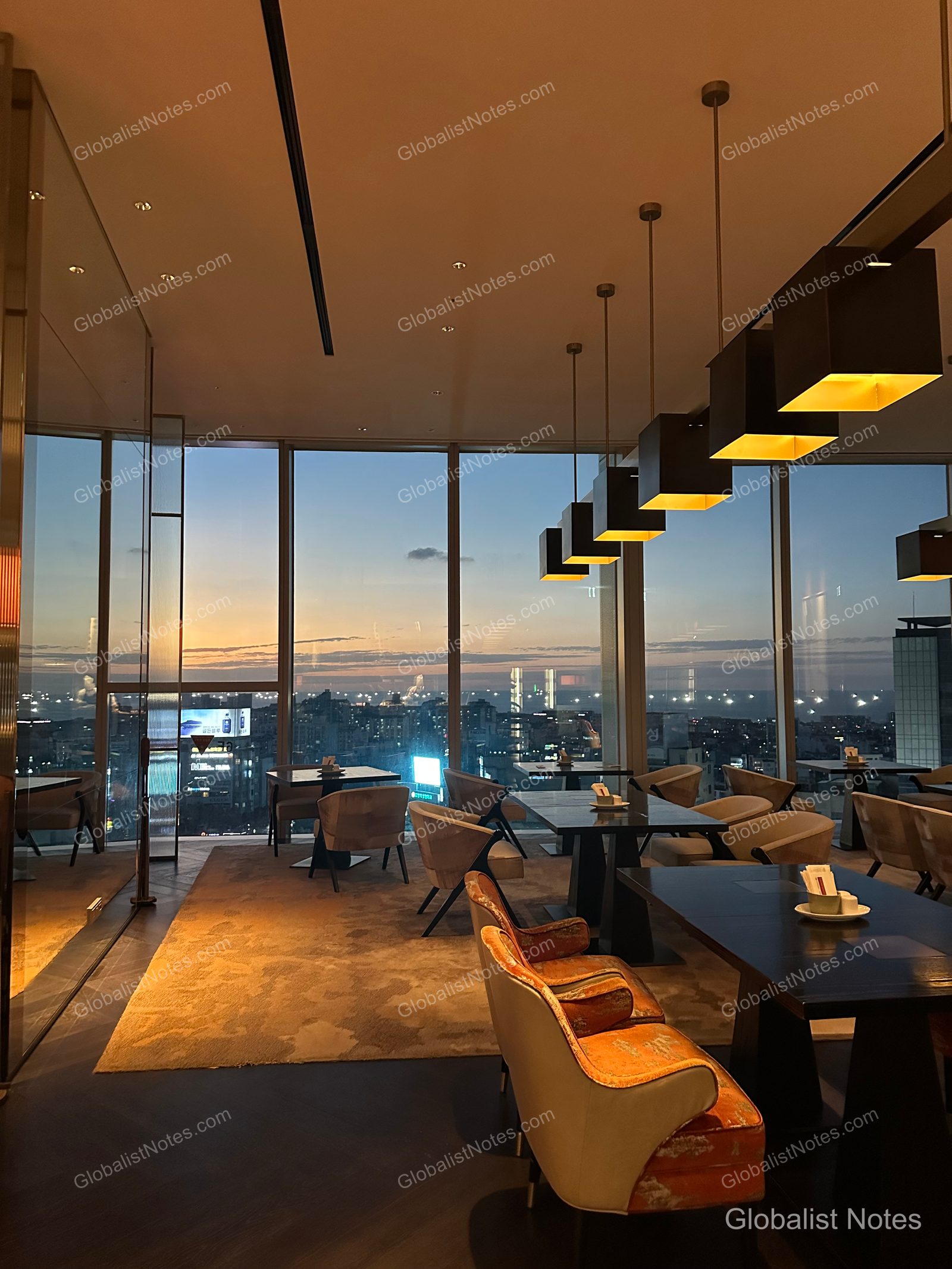 The interior of the Grand Hyatt Jeju lounge, featuring neatly arranged tables and chairs under square ceiling lights, with a view of the Jeju city skyline at sunset through floor-to-ceiling windows.