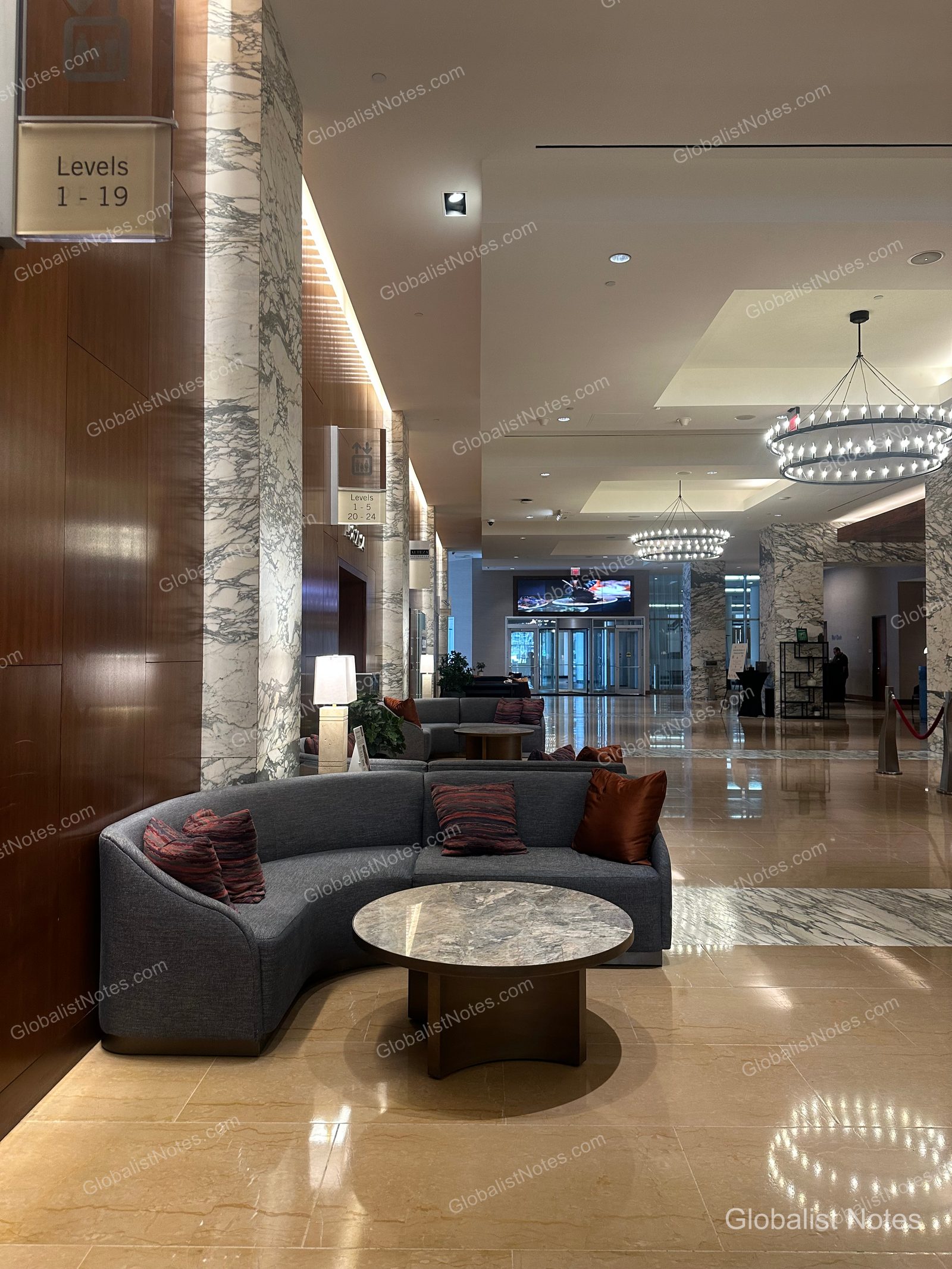 Spacious lobby of the Grand Hyatt San Antonio River Walk. Curved sofas and round tables are arranged along walls finished with marble columns and wood paneling, with a modern chandelier overhead and elevator floor signage visible.