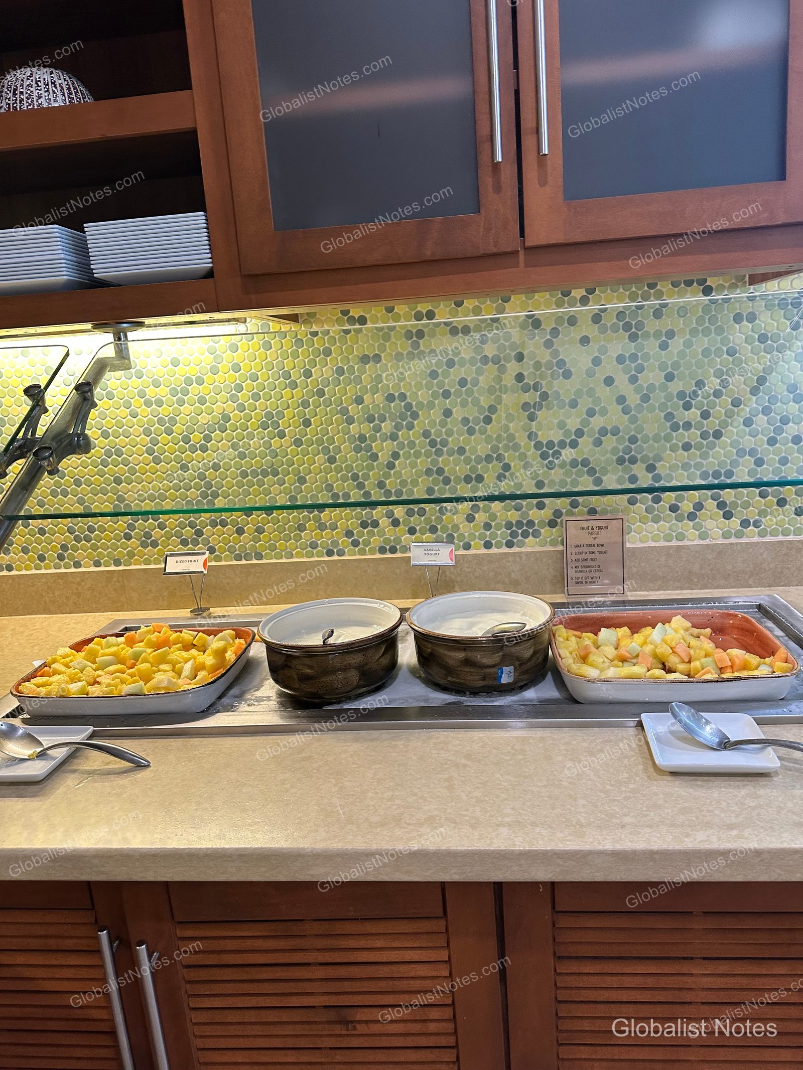 Breakfast buffet station at Hyatt Place Waikiki Beach featuring bowls of cubed fruit and yogurt, set against a green and yellow tiled wall with white plates stored on wooden shelving above.