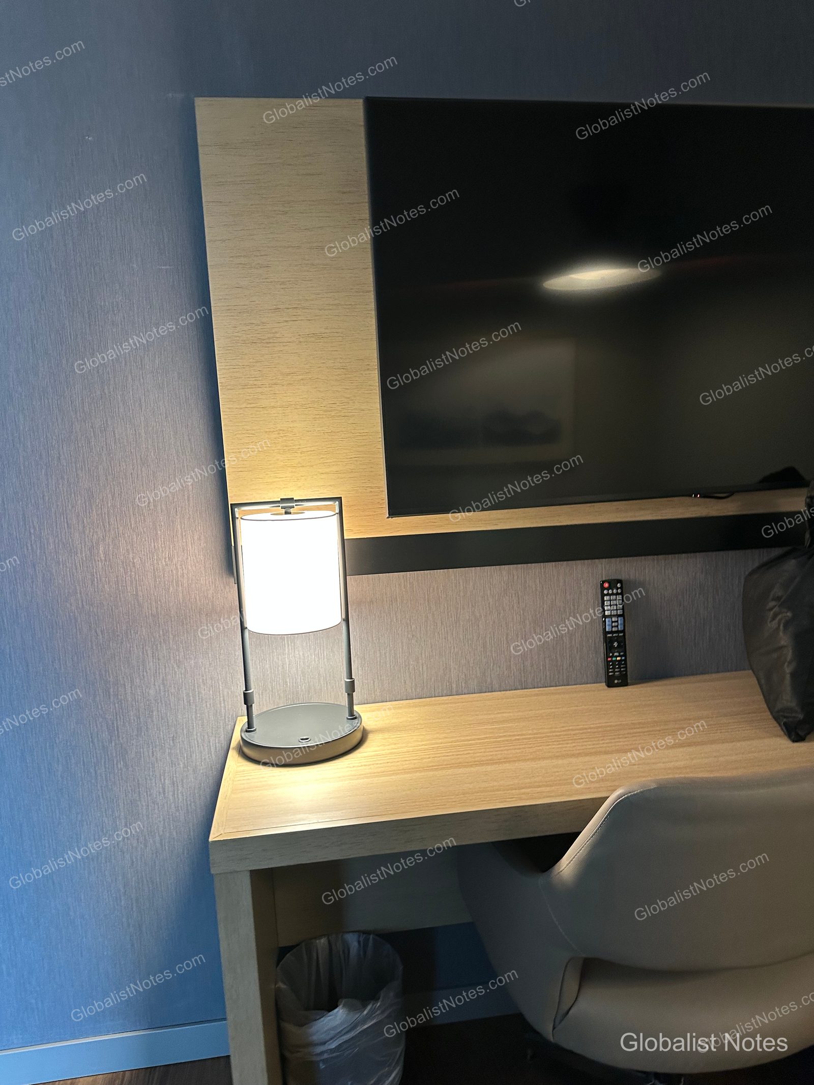Hyatt Place Bozeman Yellowstone Airportのジュニアスイートルームのワークデスク。明るい木目調のデスクにライトとリモコンが置かれ、壁には薄型テレビ、手前にはグレーの椅子が配置されています。