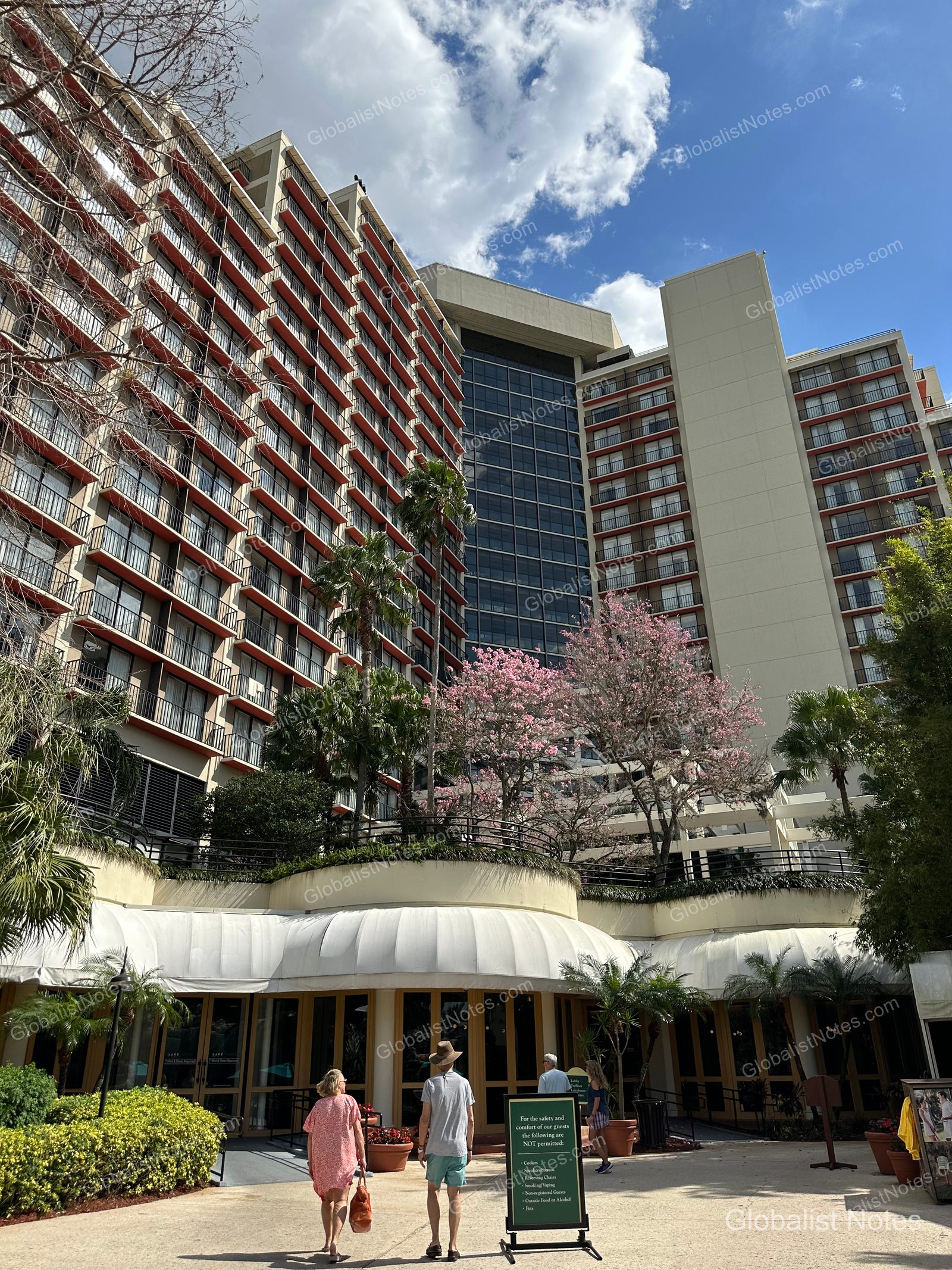 The exterior and entrance of Hyatt Regency Grand Cypress in Orlando, featuring a large building with tiered red balconies, flowering trees, palm trees, and guests walking toward the entrance.