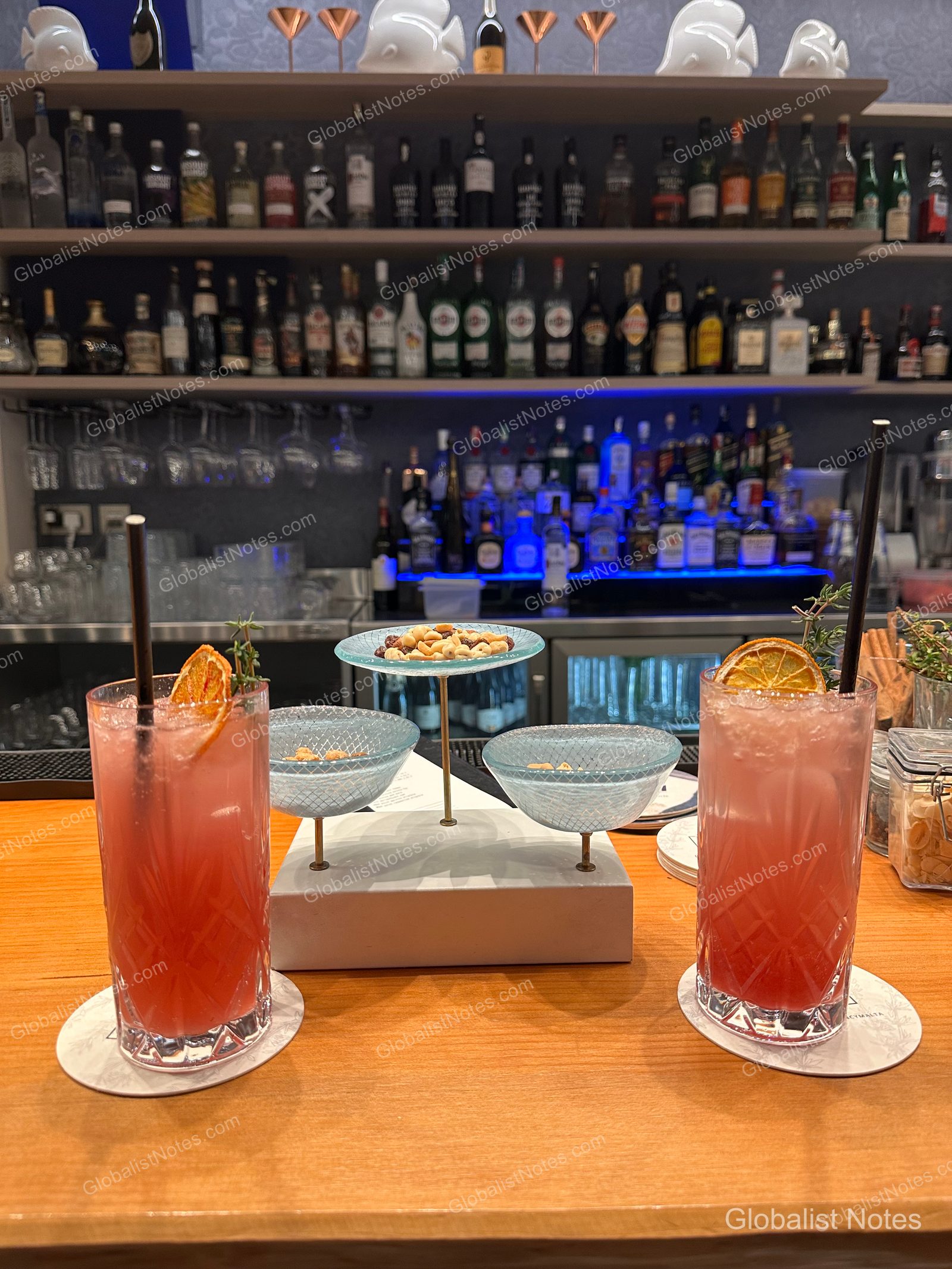 Two pink cocktails and a three-tiered dish of nuts on the bar counter at Hyatt Regency Malta, with a backlit shelf of liquor bottles in the background.