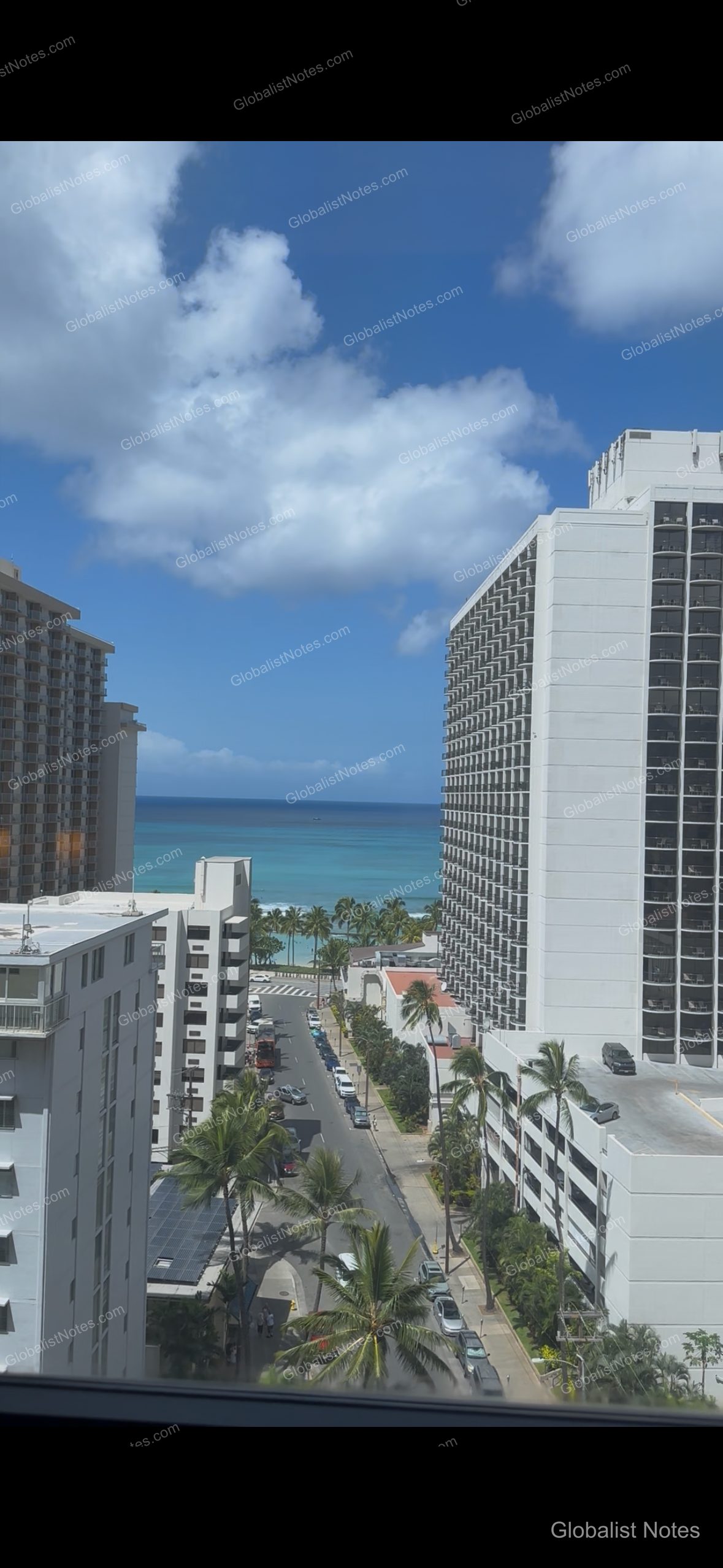View from a guest room at Hyatt Place Waikiki Beach in Honolulu, Hawaii, showing a glimpse of the blue ocean between buildings, palm-lined streets, and the urban Waikiki landscape under a clear sky.