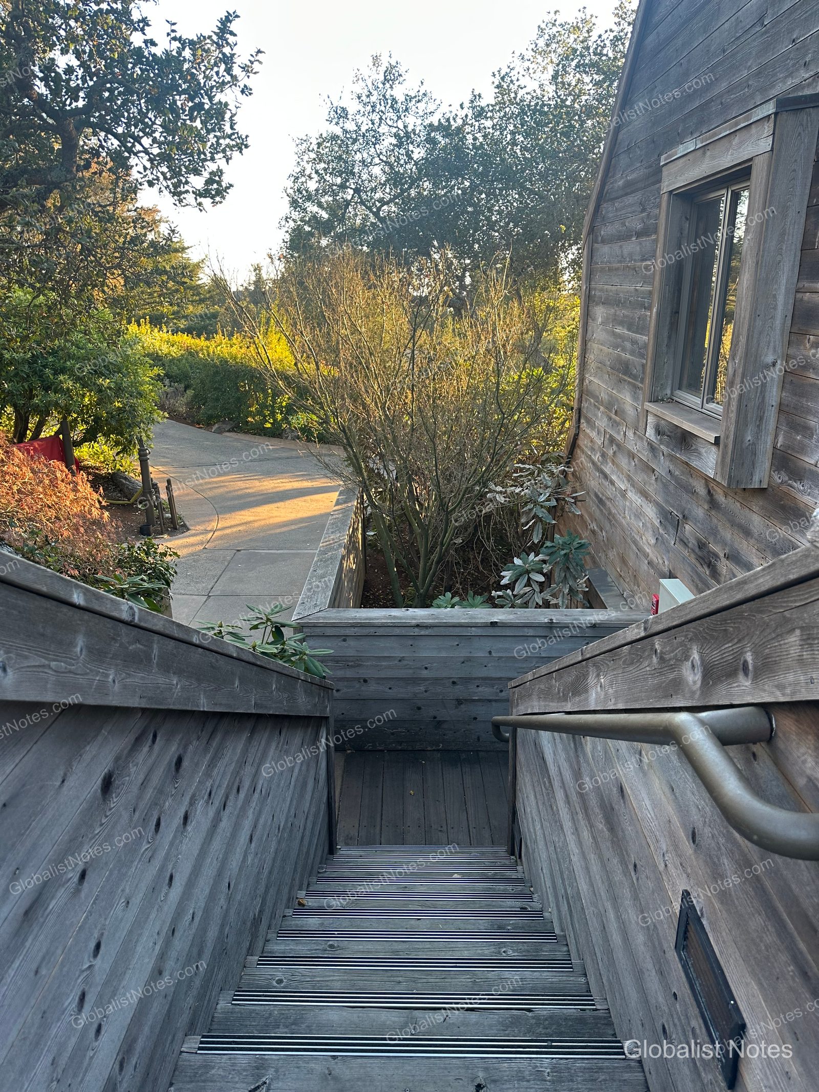 A view from the outdoor wooden stairs at Alila Ventana Big Sur, showing the wood-paneled guest room building on the right and a sunlit path and garden on the left.
