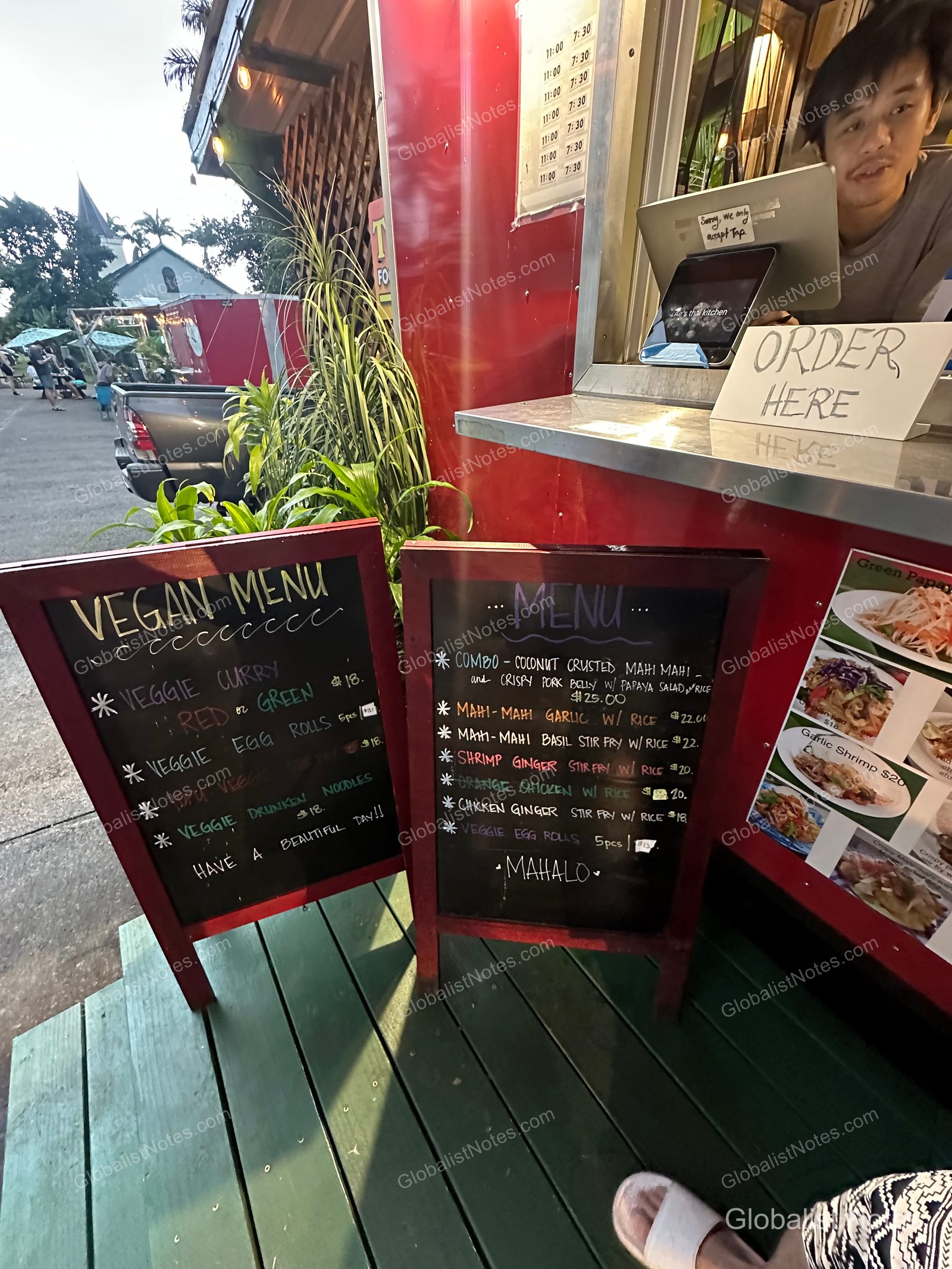Two black sandwich boards displaying vegan and standard menu options stand in front of a food truck near Hana-Maui Resort, with a staff member assisting a customer at the order window.