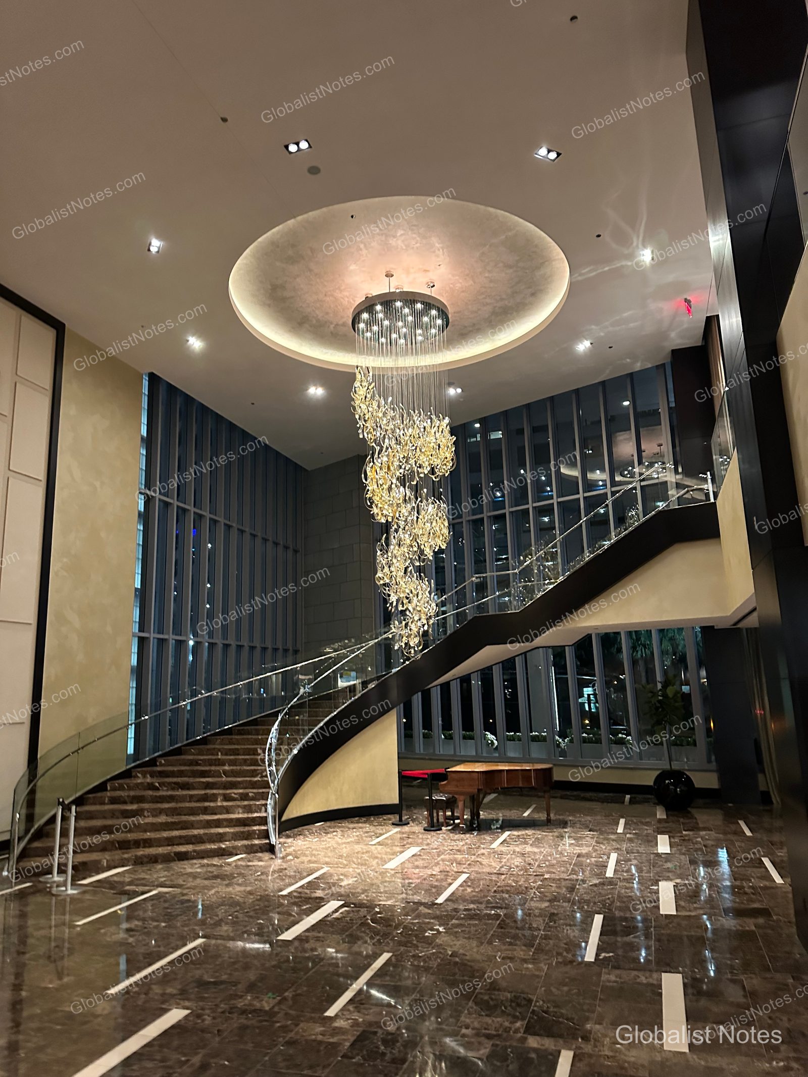 Lobby view at Hyatt Regency New Orleans, featuring a high ceiling with a central spiral crystal chandelier, a curved staircase, and a grand piano. The space has marble flooring and floor-to-ceiling windows that create an open atmosphere.