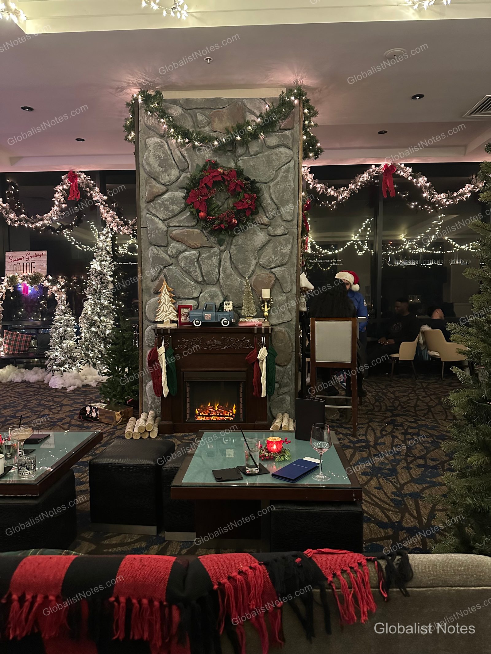 Manchester Grand Hyatt San Diego 的室內空間，石砌壁爐上方掛著聖誕花圈，周圍點綴著聖誕樹與裝飾，前方擺放的桌椅營造出溫馨的用餐氛圍。