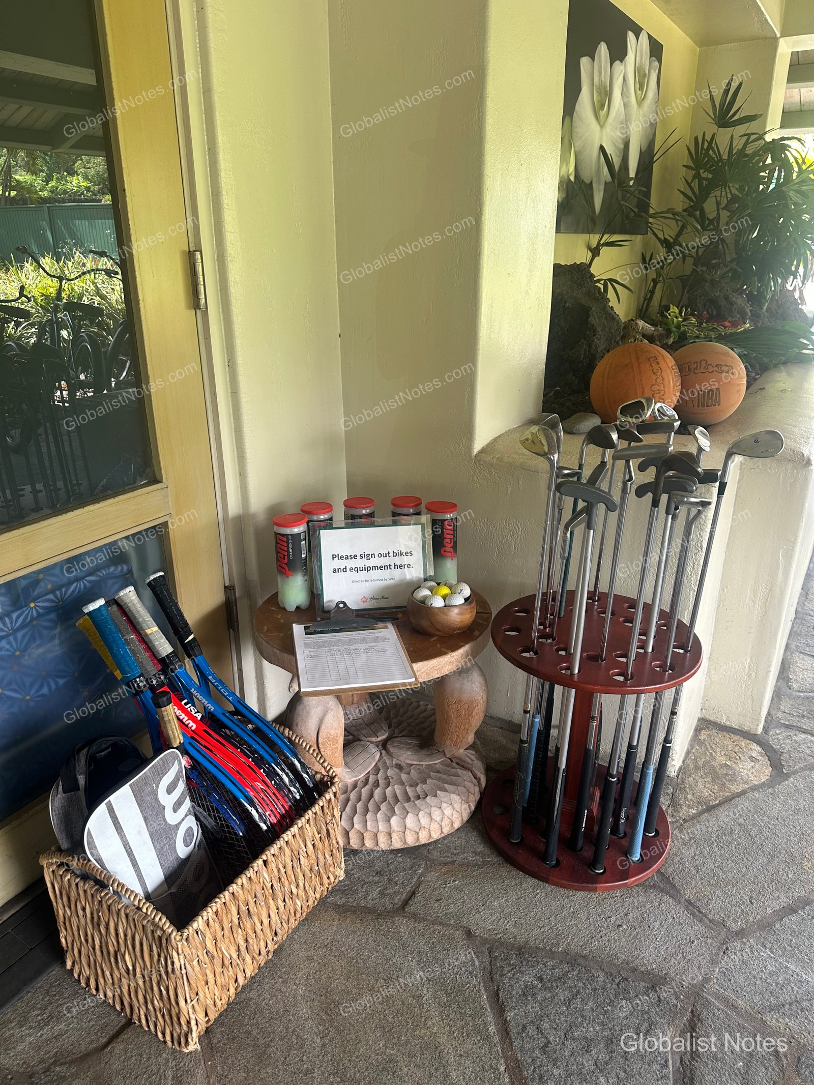 A basket of tennis rackets, a golf club rack, and basketballs are available in the outdoor area at Hana-Maui Resort. A small wooden table in the center holds a clipboard for rental sign-ups, informational notices, and a canister of tennis balls, allowing guests to use the sports equipment at their leisure.