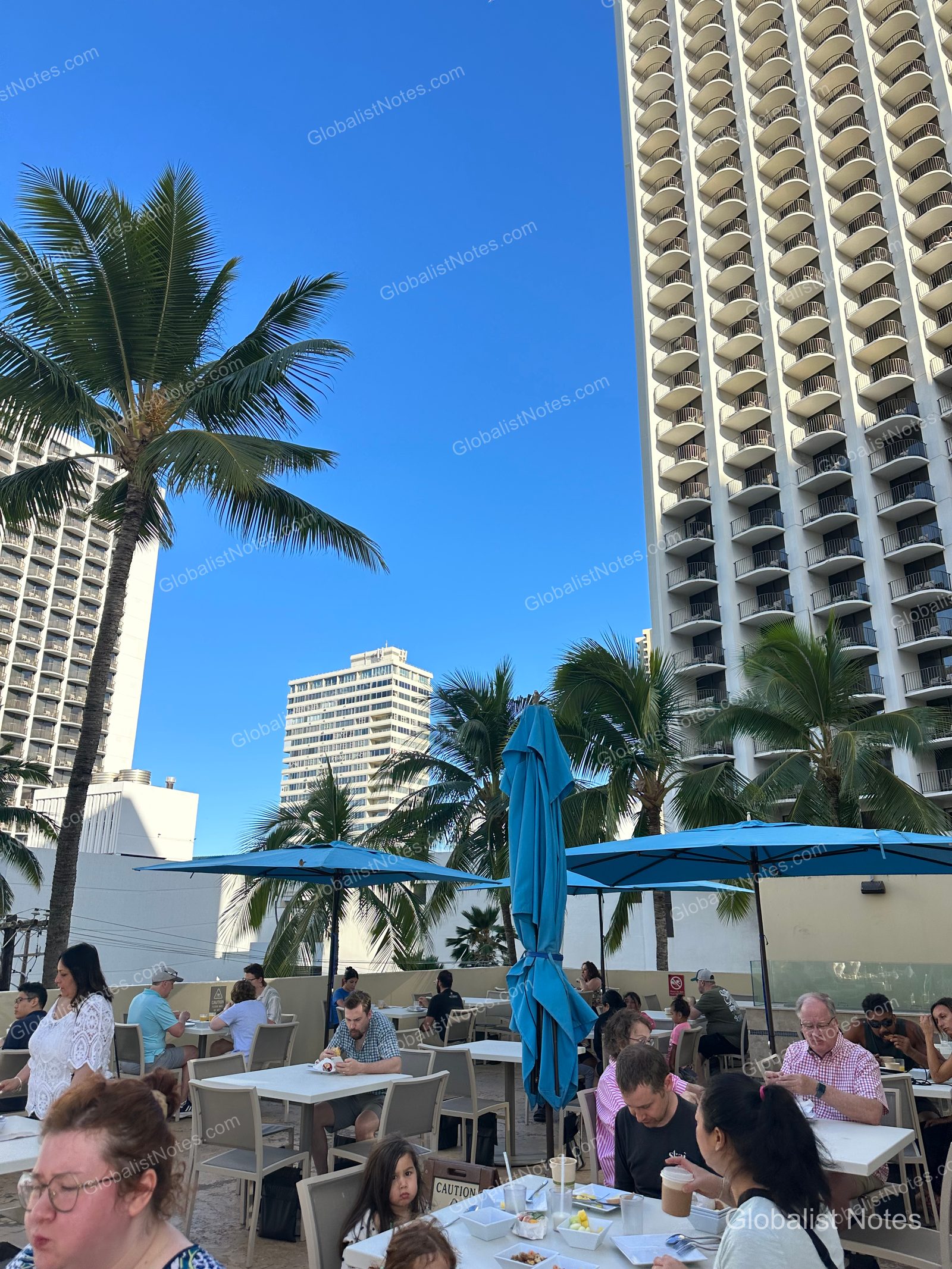 Outdoor breakfast area at Hyatt Place Waikiki Beach in Honolulu, Hawaii. Blue umbrellas are set up on a terrace lined with palm trees under a clear sky, with guests seated at tables.