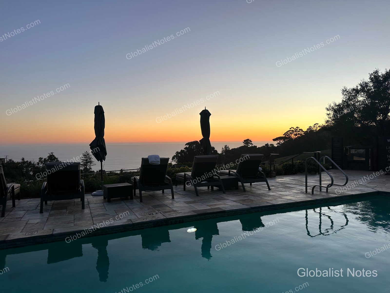 The outdoor pool deck at Alila Ventana Big Sur at sunset. Sun loungers and umbrellas are arranged with the Pacific Ocean in the background, and the orange glow of the sky reflects on the pool water.