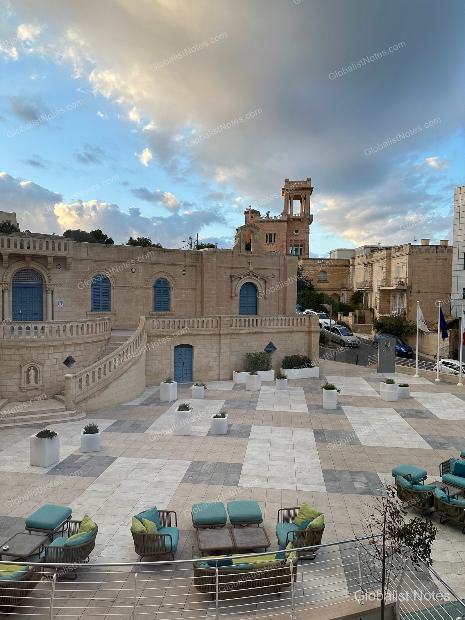View of the square from a Junior Suite balcony at Hyatt Regency Malta, overlooking antique stone buildings, a church with blue doors, and terrace seating below.