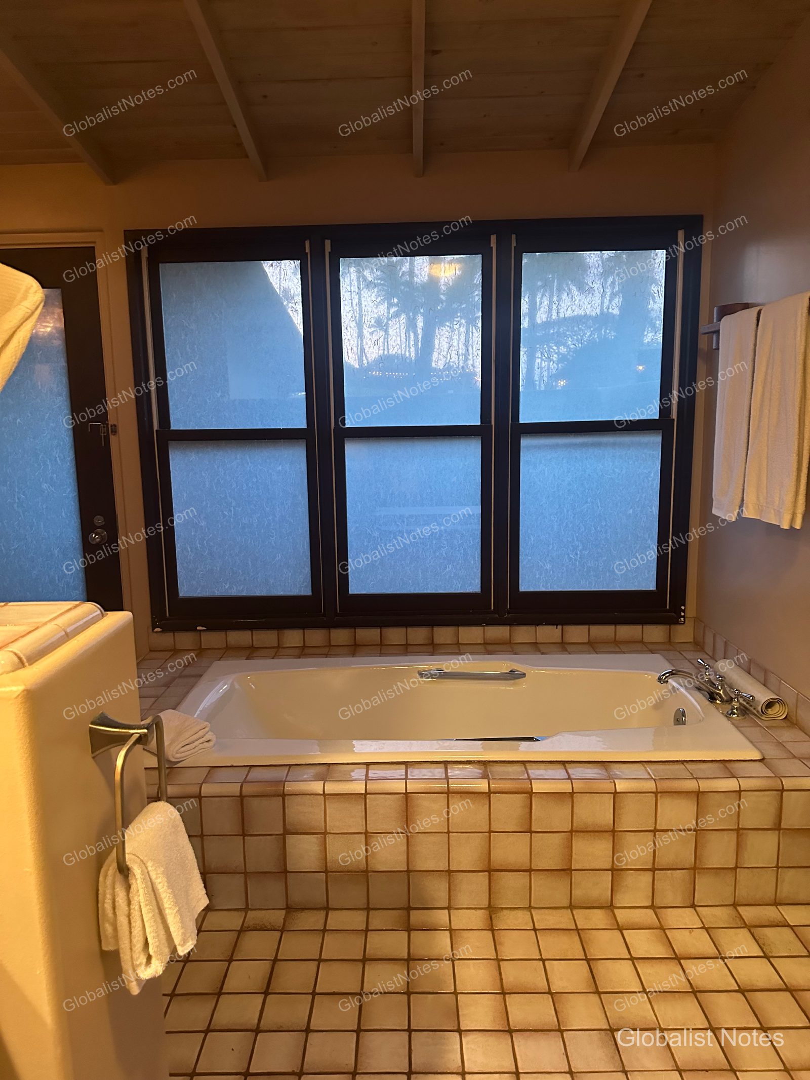 Bathroom interior of a 1 King Bed Ocean View Bungalow at Hana-Maui Resort. A spacious tiled soaking tub is positioned by the window, which overlooks Maui’s palm trees and natural landscape. White towels are hung beside the tub, and the interior features a calm, warm color palette.