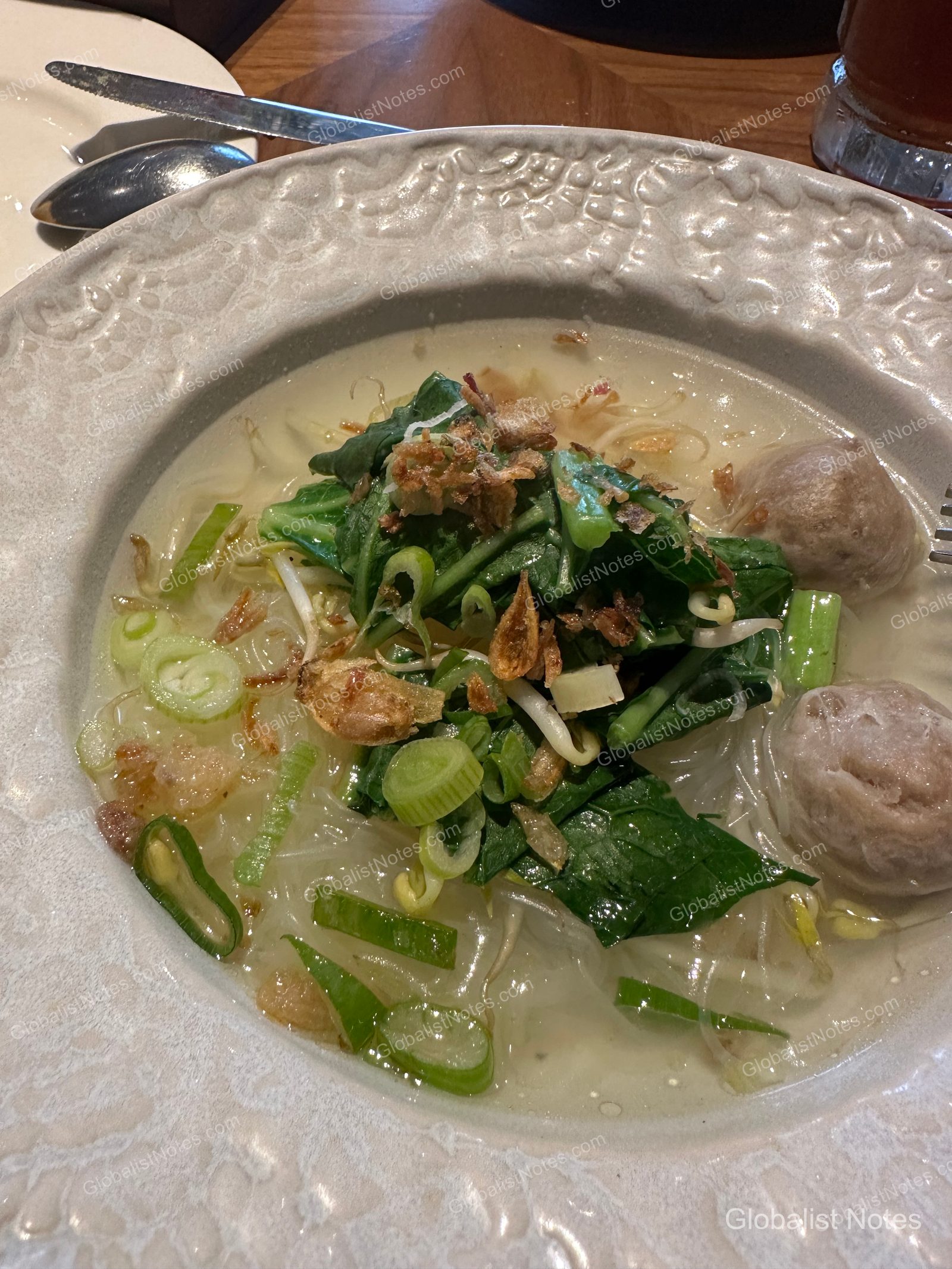 A bowl of Bakso, an Indonesian meatball noodle soup, served at the Grand Hyatt Jakarta breakfast buffet. The soup is topped with sliced scallions, bean sprouts, fried shallots, and leafy greens.