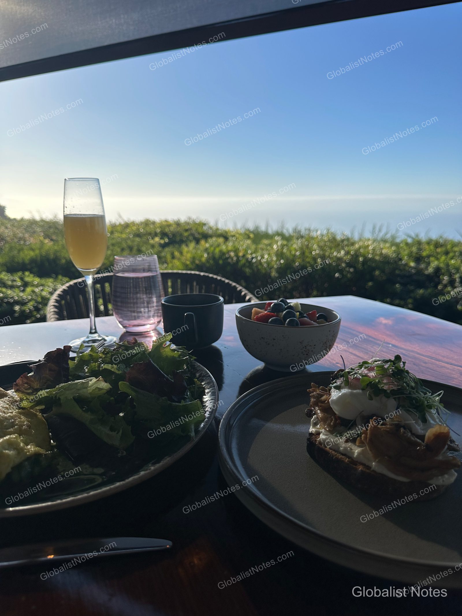 Breakfast spread on an outdoor terrace table at Alila Ventana Big Sur, including salad, eggs Benedict, a fruit bowl, and champagne glasses, with the blue sea and horizon of Big Sur in the background.
