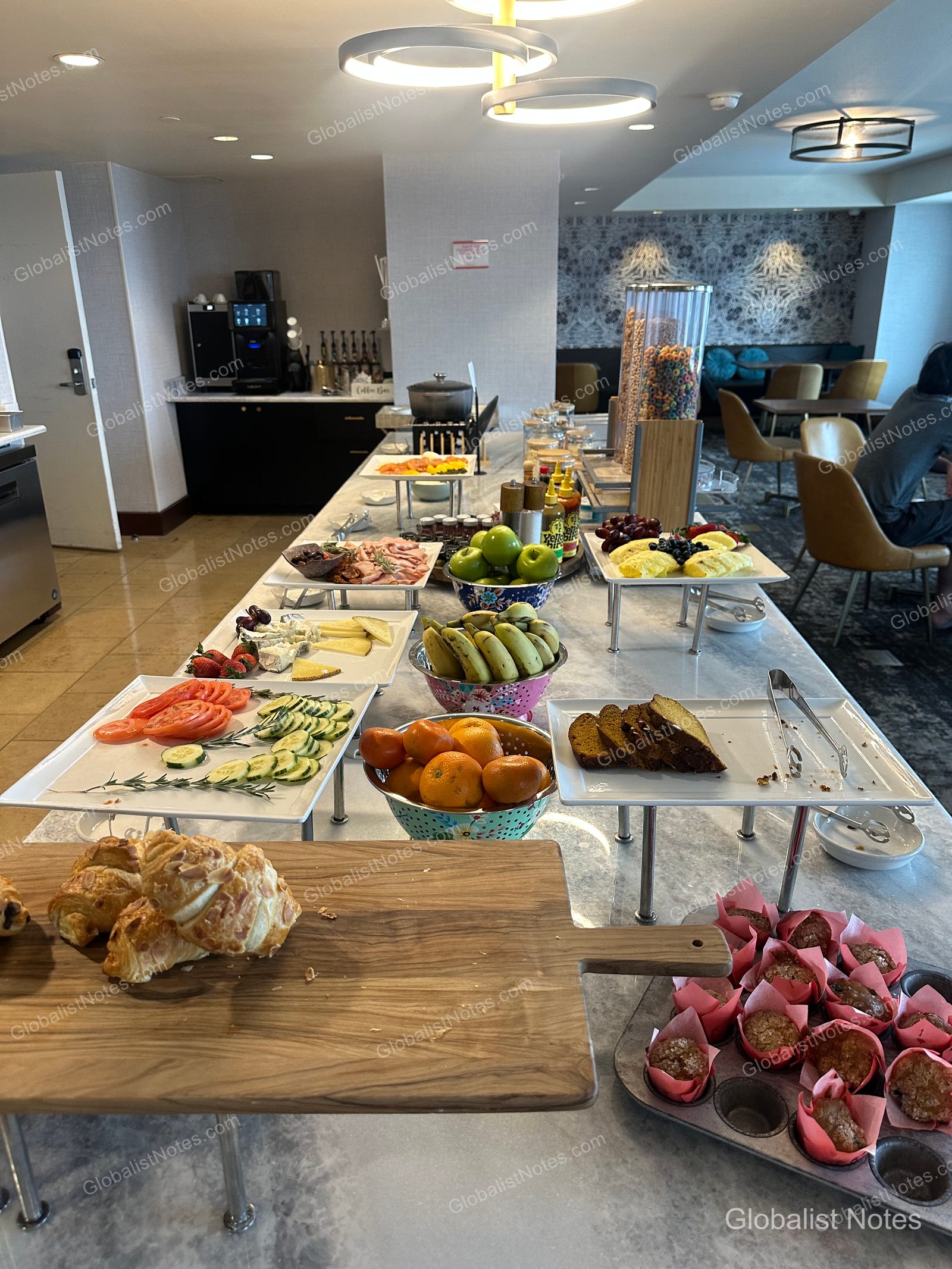 A long marble table set with the breakfast buffet at Grand Hyatt San Antonio River Walk. The spread features fresh fruit, sliced vegetables, cheeses, meats, croissants, and muffins, with coffee machines and seating visible in the background.