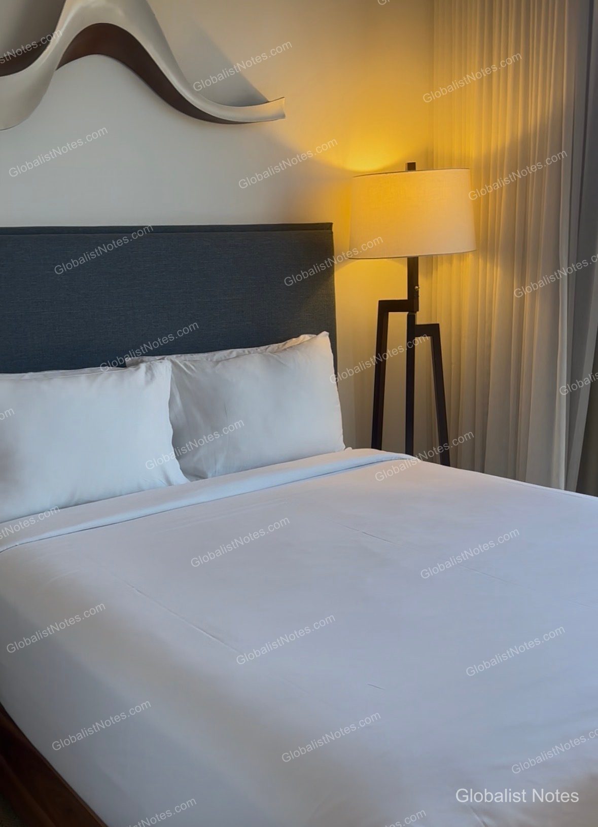 A view of the bedside area in a Hyatt Centric Waikiki Beach guest room, featuring a dark gray fabric headboard, crisp white bedding, and a warm-toned floor lamp. A curved decorative element is mounted on the wall above the bed.