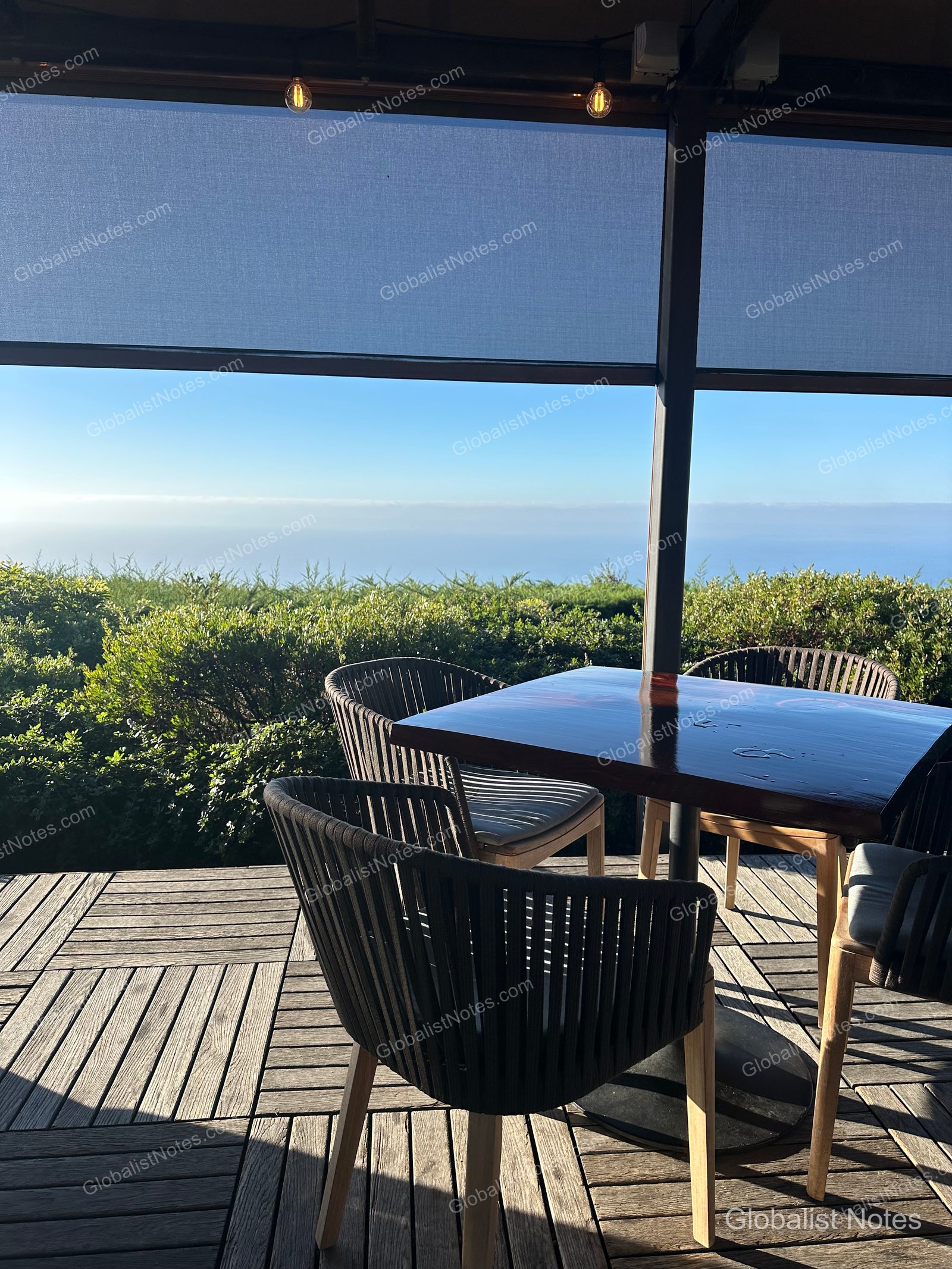 Outdoor terrace dining at Alila Ventana Big Sur with tables and chairs on a wooden deck overlooking the ocean horizon under a retractable awning in the morning sun.