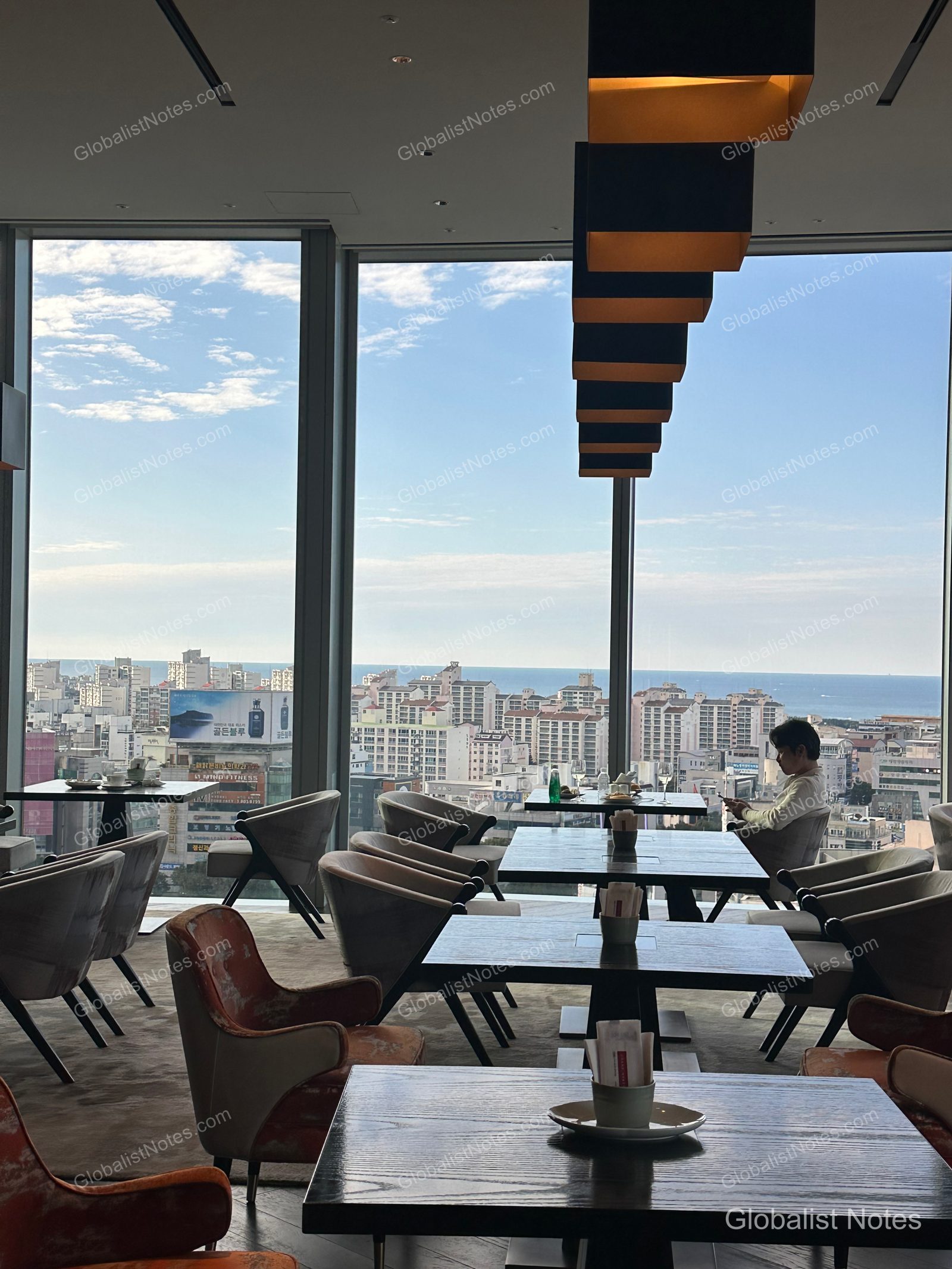 Interior view of the lounge at Grand Hyatt Jeju, featuring tables and chairs neatly arranged beneath rectangular pendant lighting. Floor-to-ceiling windows offer a view of the Jeju city skyline and the distant ocean.