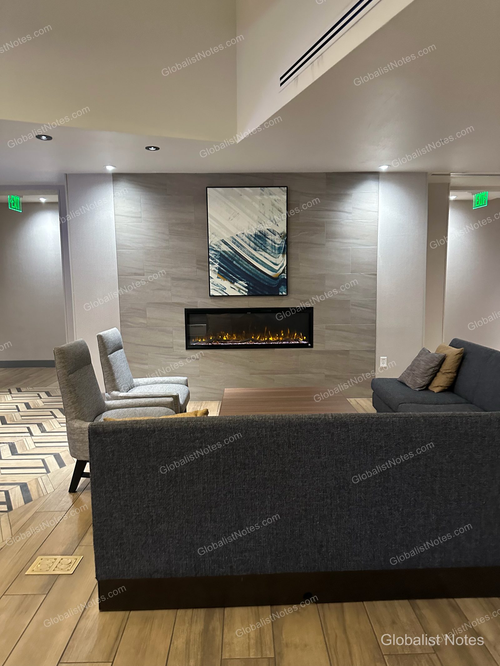 Hyatt Place Flagstaffのロビーにある休憩スペース。壁面中央に埋め込み式の暖炉と抽象画があり、その前にグレーのソファと1人掛けチェア、木製テーブルが置かれています。床は明るい木目調のタイル張りです。
