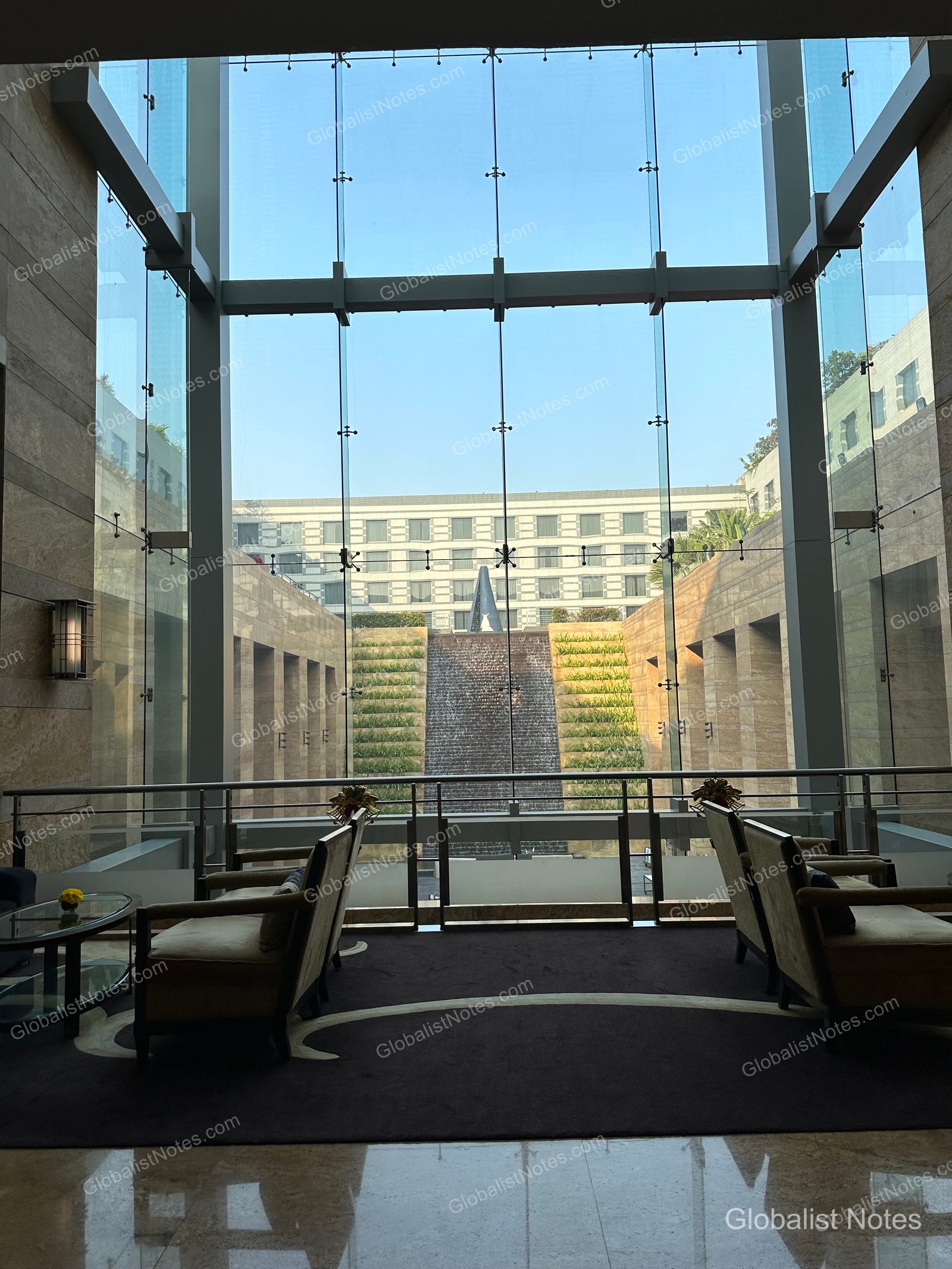The lobby lounge at Grand Hyatt Mumbai, featuring floor-to-ceiling windows overlooking the tiered waterfall and gardens. Symmetrical seating is arranged by the windows, where natural light creates a calm atmosphere.