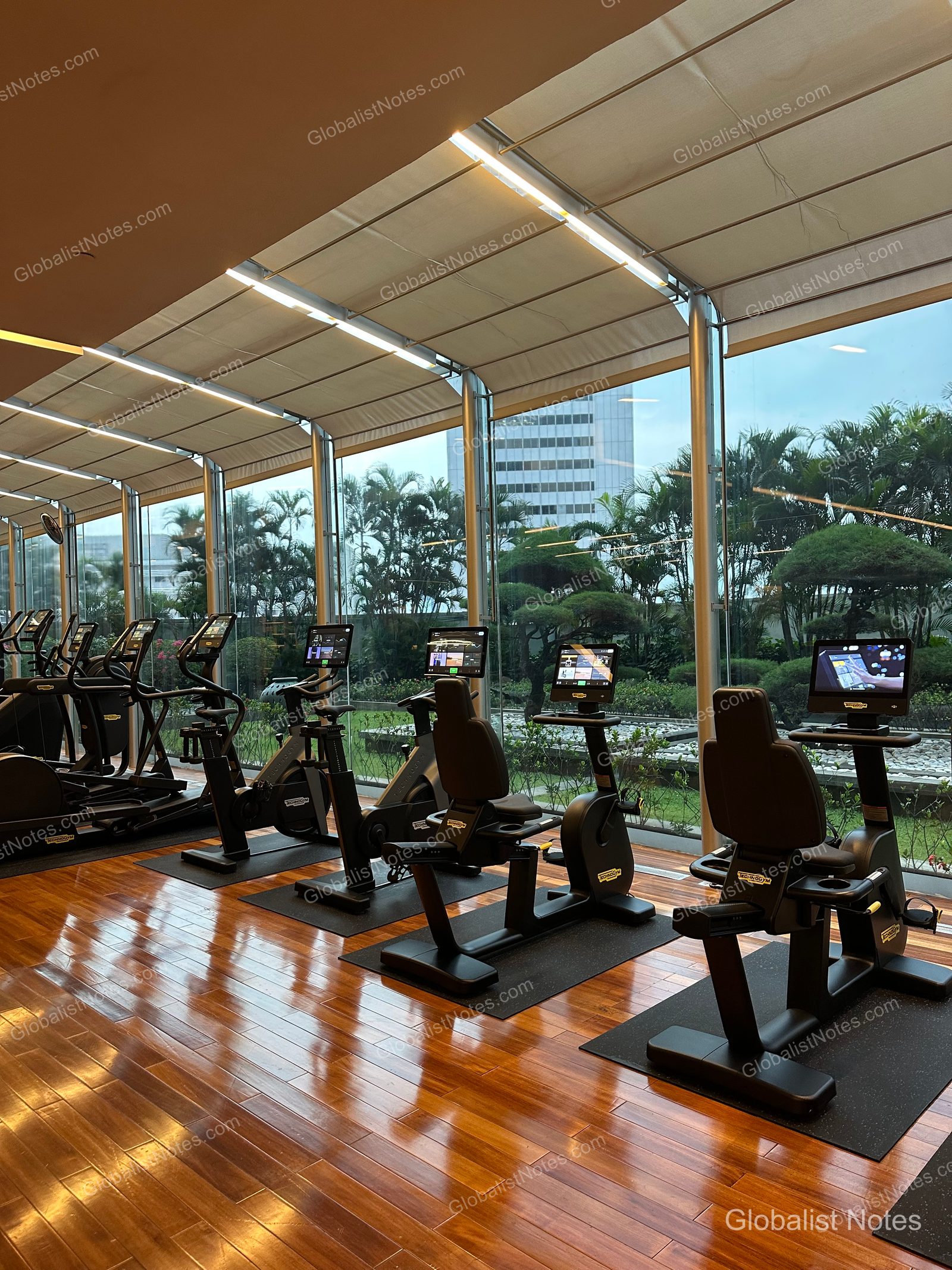 Interior of the fitness center at Grand Hyatt Jakarta. Technogym stationary bikes and treadmills are lined up along floor-to-ceiling windows overlooking the garden and city skyline. The floor is finished with polished wood.