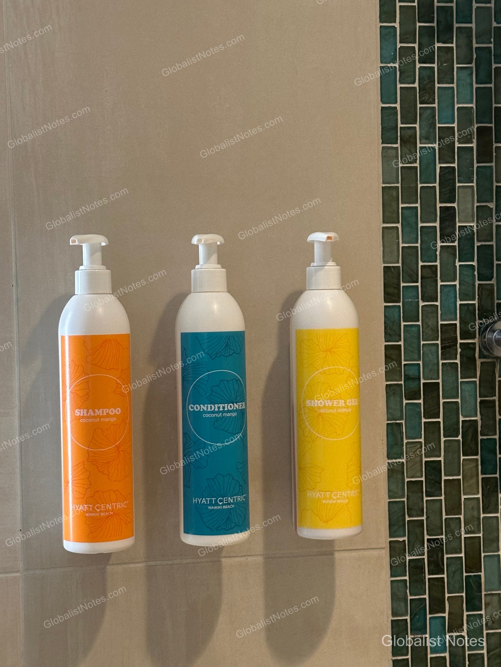 Wall-mounted pump dispensers for shampoo, conditioner, and body wash in a Hyatt Centric Waikiki Beach guest room bathroom. The containers are arranged in a row—orange for shampoo, blue for conditioner, and yellow for shower gel—against a teal tiled wall.