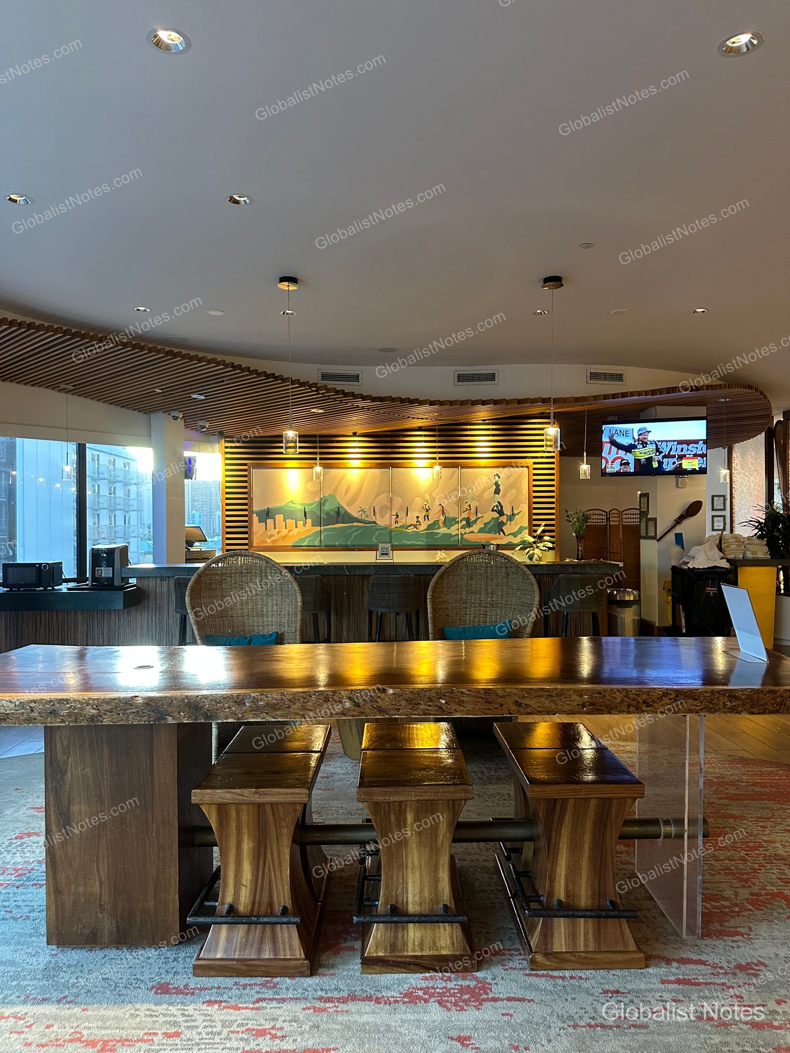 Lobby lounge at Hyatt Centric Waikiki Beach, featuring a long wooden communal table with matching stools in the center. The back wall behind the bar counter is decorated with Hawaiian landscape art and a television, while the ceiling is accented with wave-like wooden slats under warm ambient lighting.