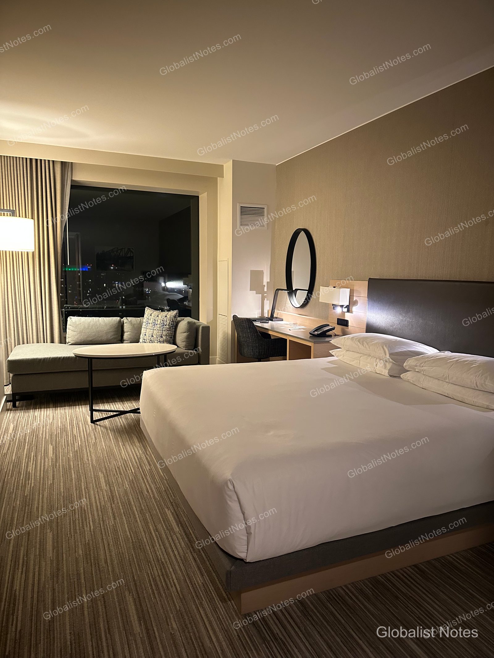 Interior of a standard king room at Hyatt Regency Seattle. A king bed with white linens is centered in the room, with a gray sofa and round table placed by the window. A desk and an oval mirror are on the wall, and the Seattle city skyline is visible through the window at night.