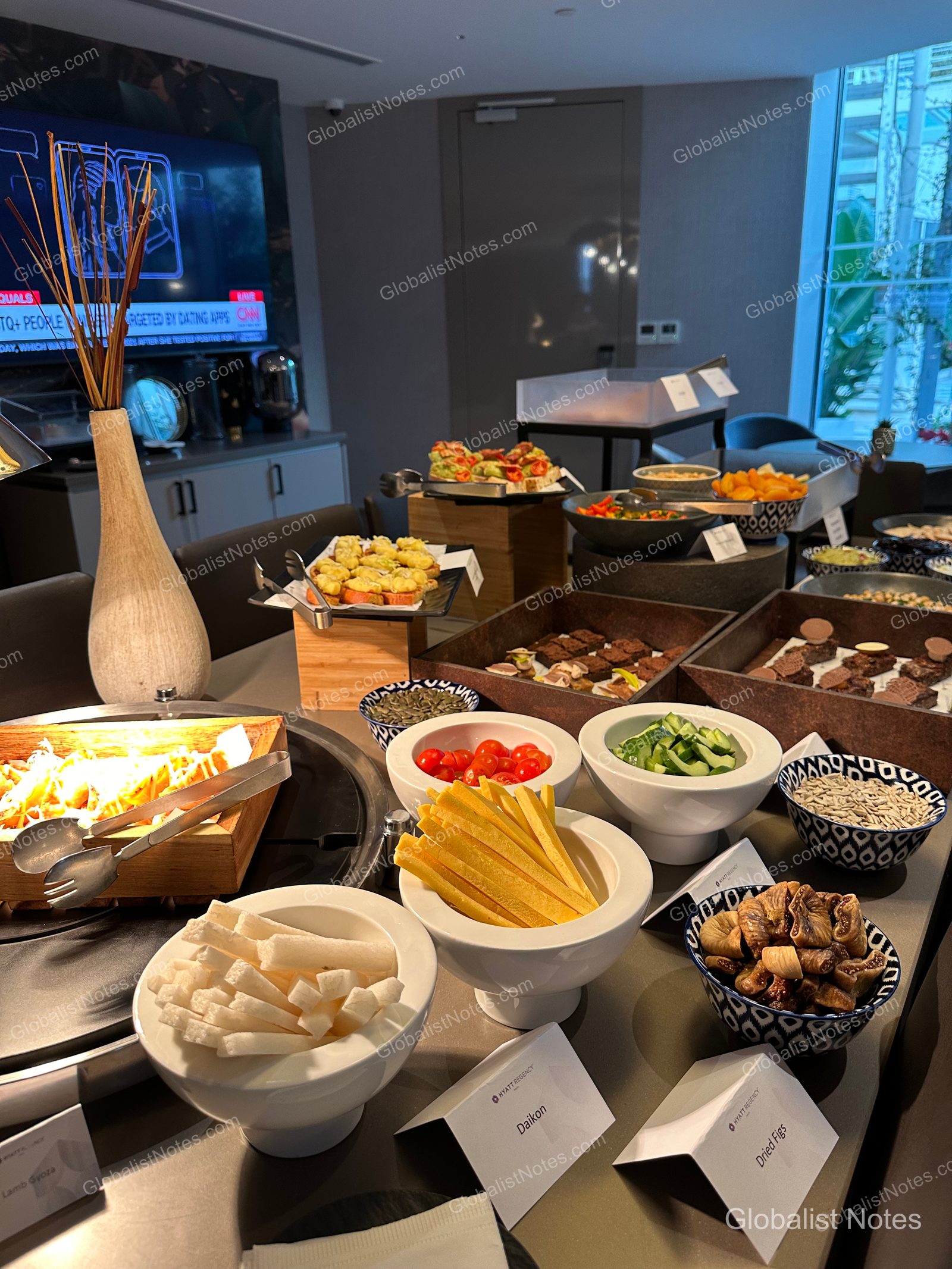 The breakfast buffet at Hyatt Regency Malta featuring bowls of fresh vegetables and fruits—including daikon, dried figs, cucumbers, and cherry tomatoes—with trays of bread and hot dishes arranged in the background.